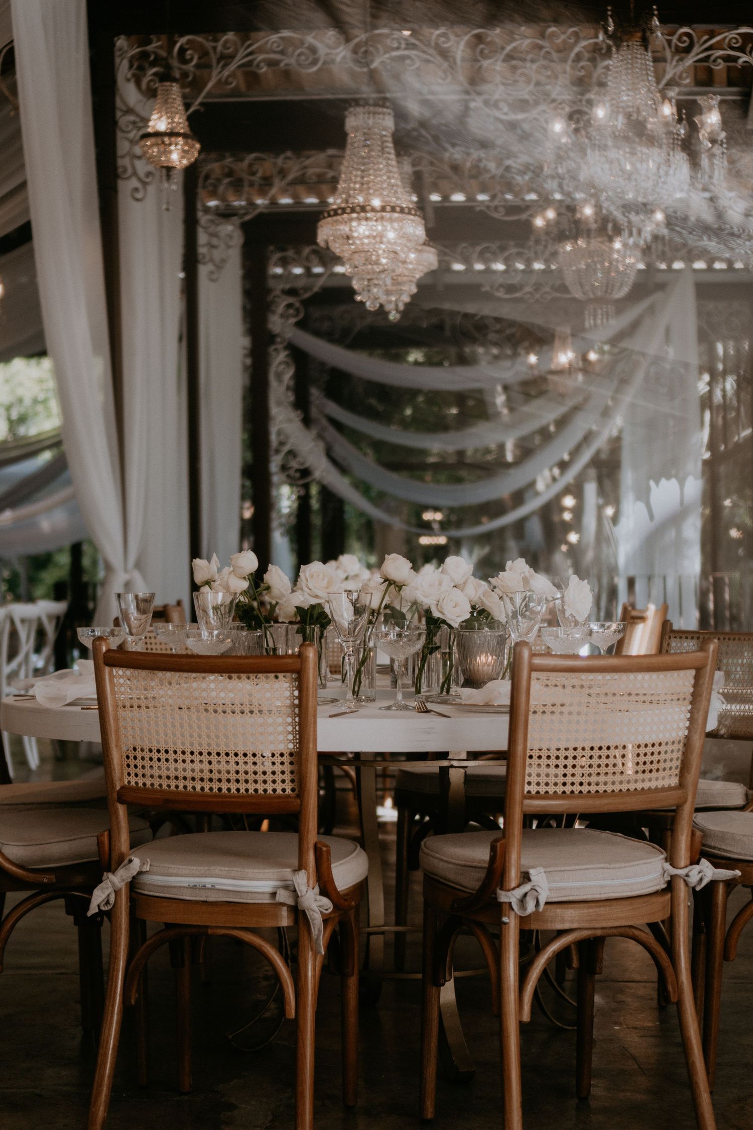 Romantic dining setup with crystal chandeliers, draped fabric, and cane-back chairs.