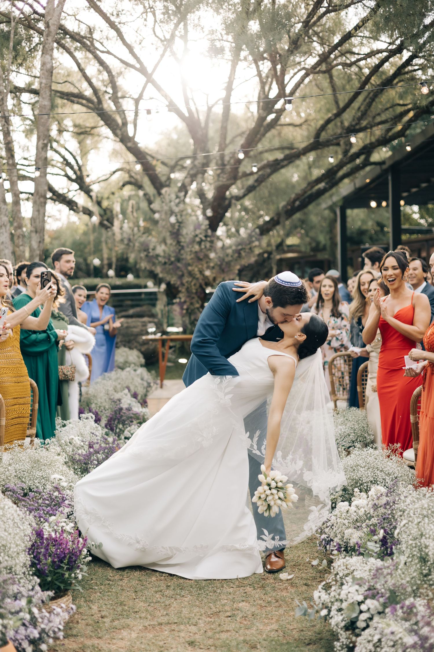 A romantic wedding couple performs dance dips amidst purple wildflowers and trees with their wedding party looking on.