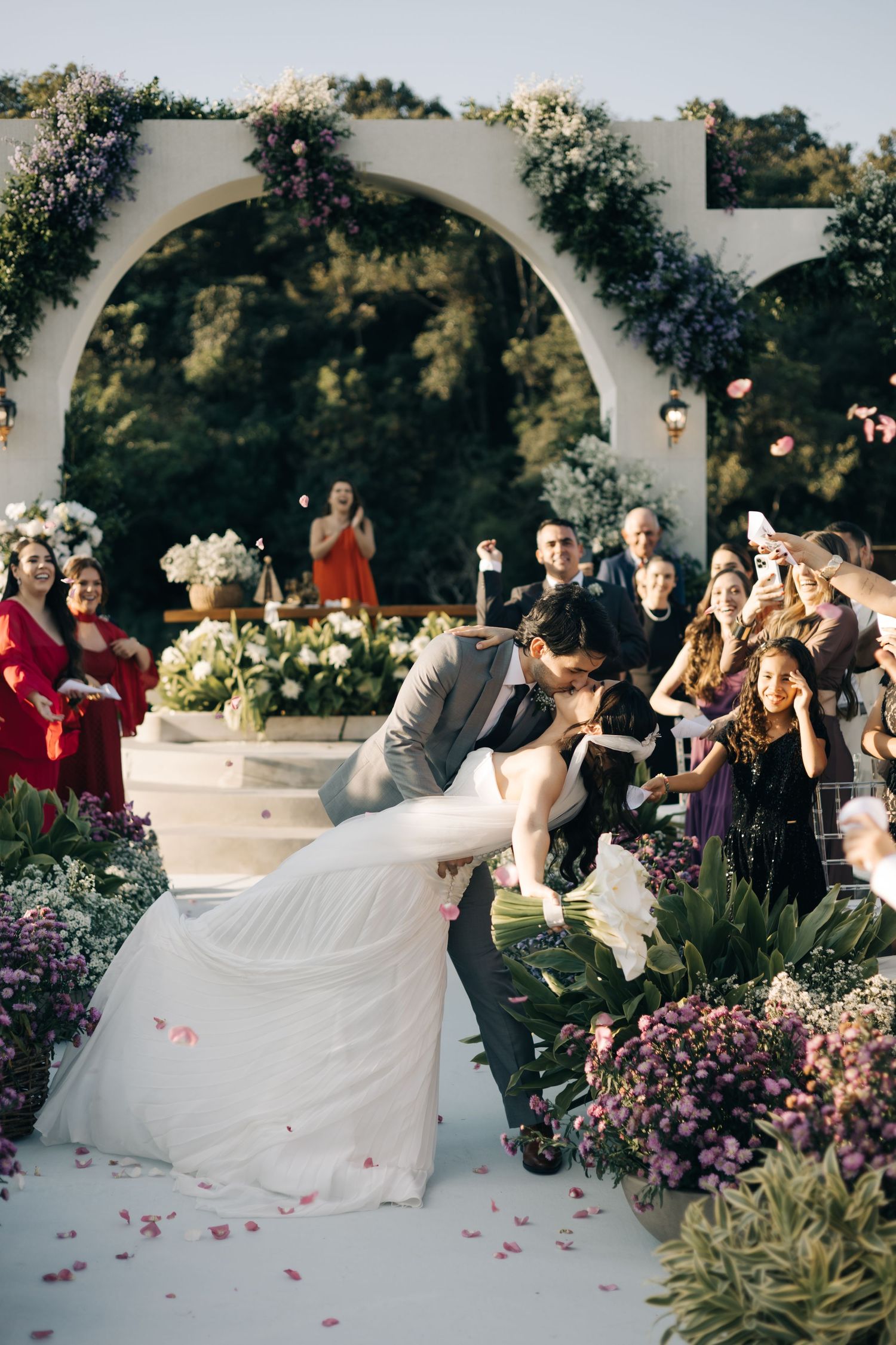 A wedding couple and guests enjoy a joyful moment in a garden setting under white arches with purple flowers.