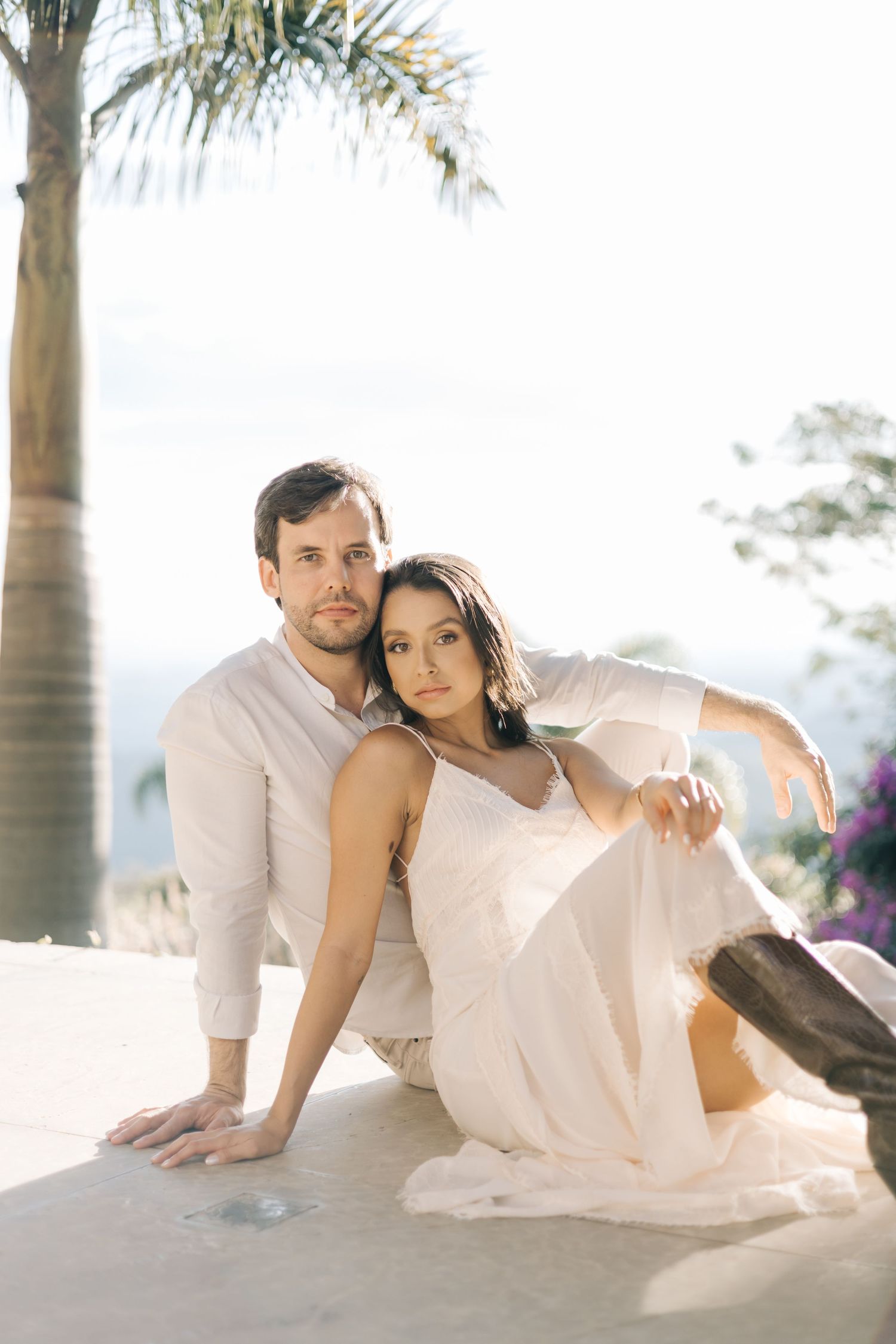 A romantic couple sits outdoors in elegant white attire with a palm tree and natural sunlight in the background.