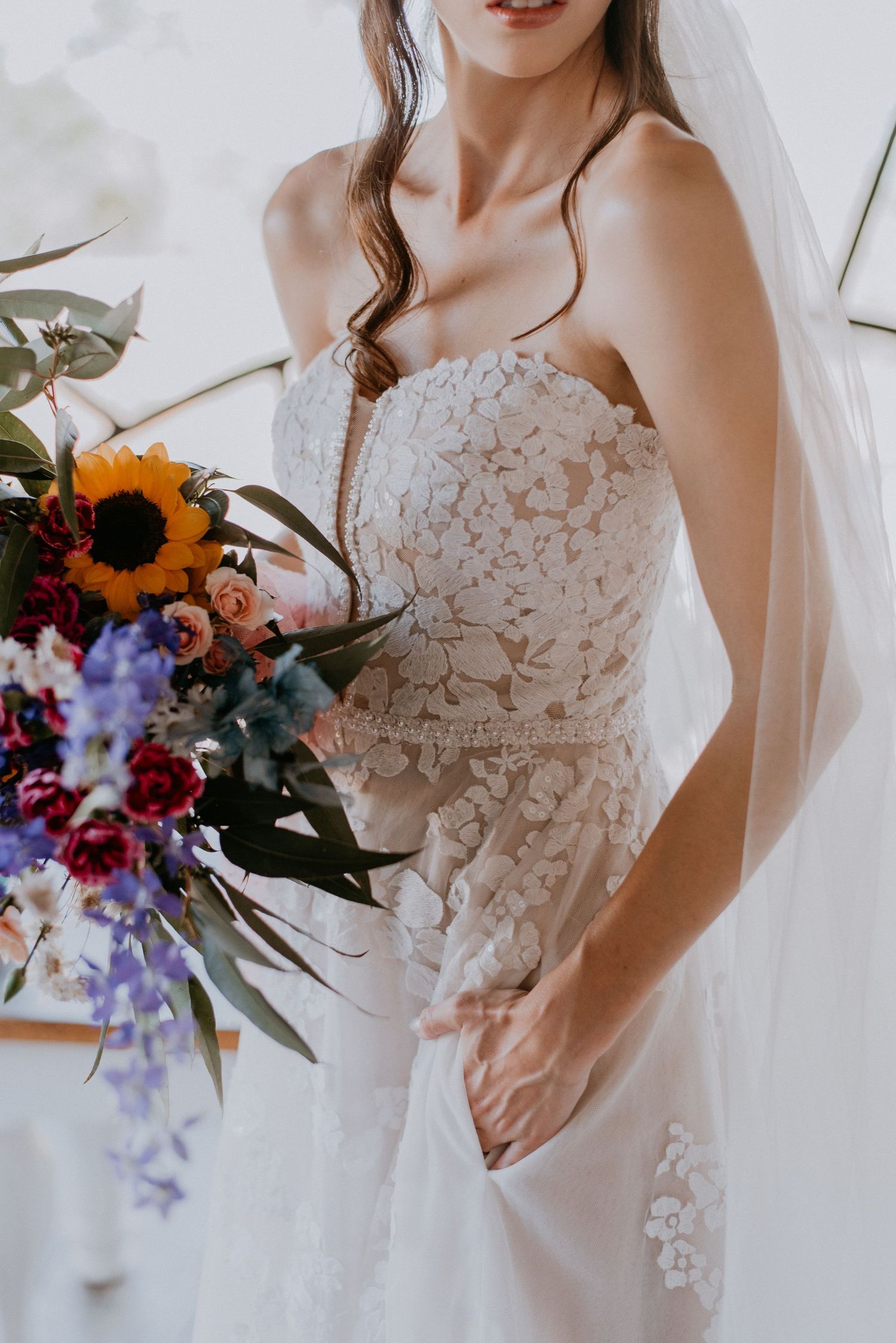 Close-up of intricate lace wedding dress with colorful sunflower and wildflower bouquet.