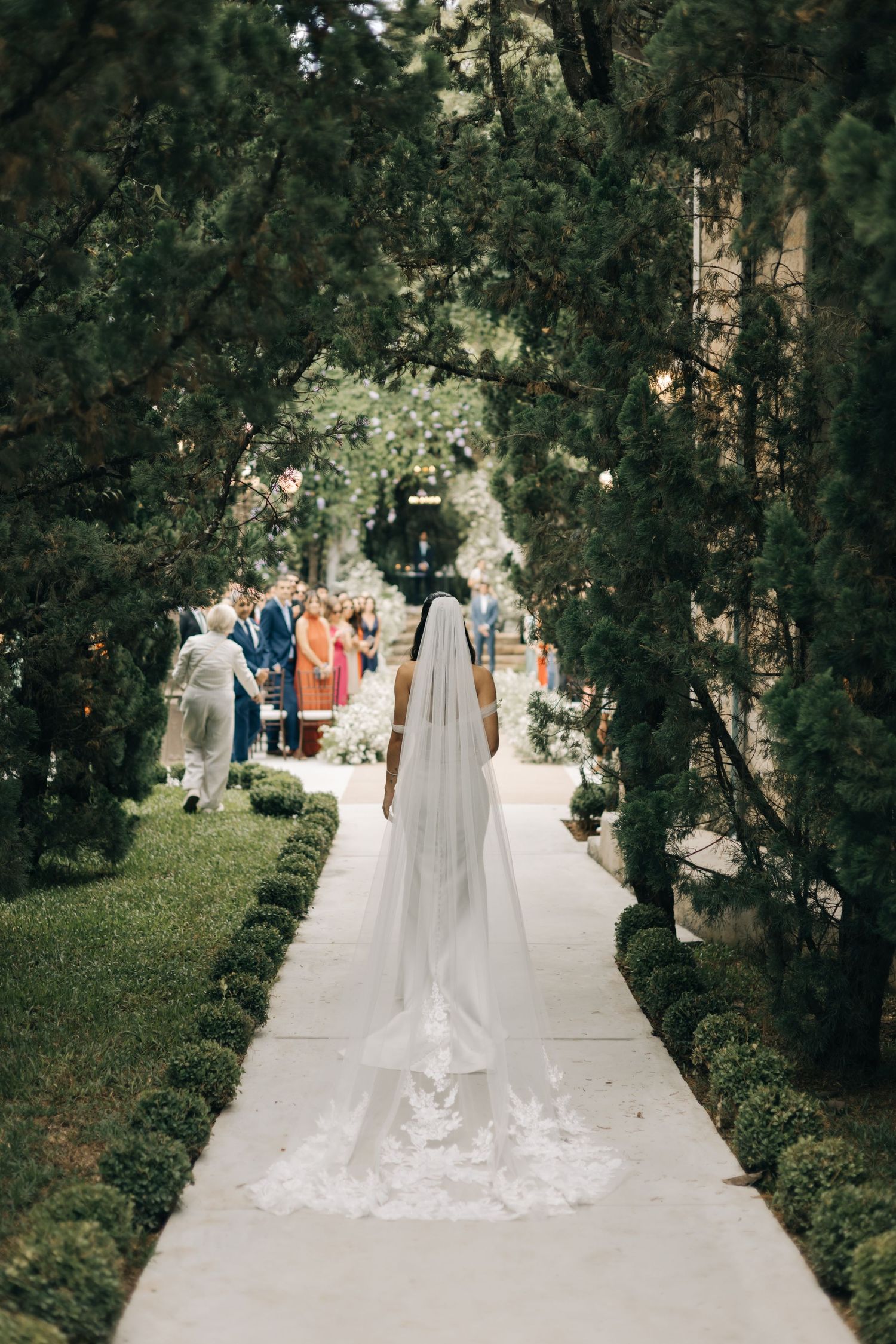 Long bridal veil trailing down a garden pathway with wedding guests visible in the background.