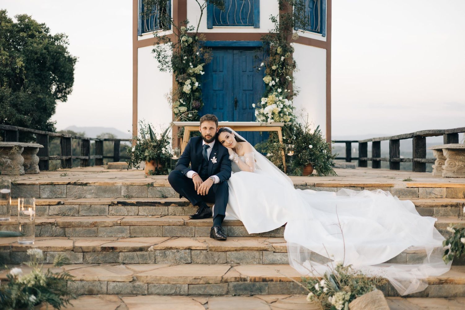 A couple in wedding attire sits together on stone steps decorated with flowers in front of a blue chapel door.