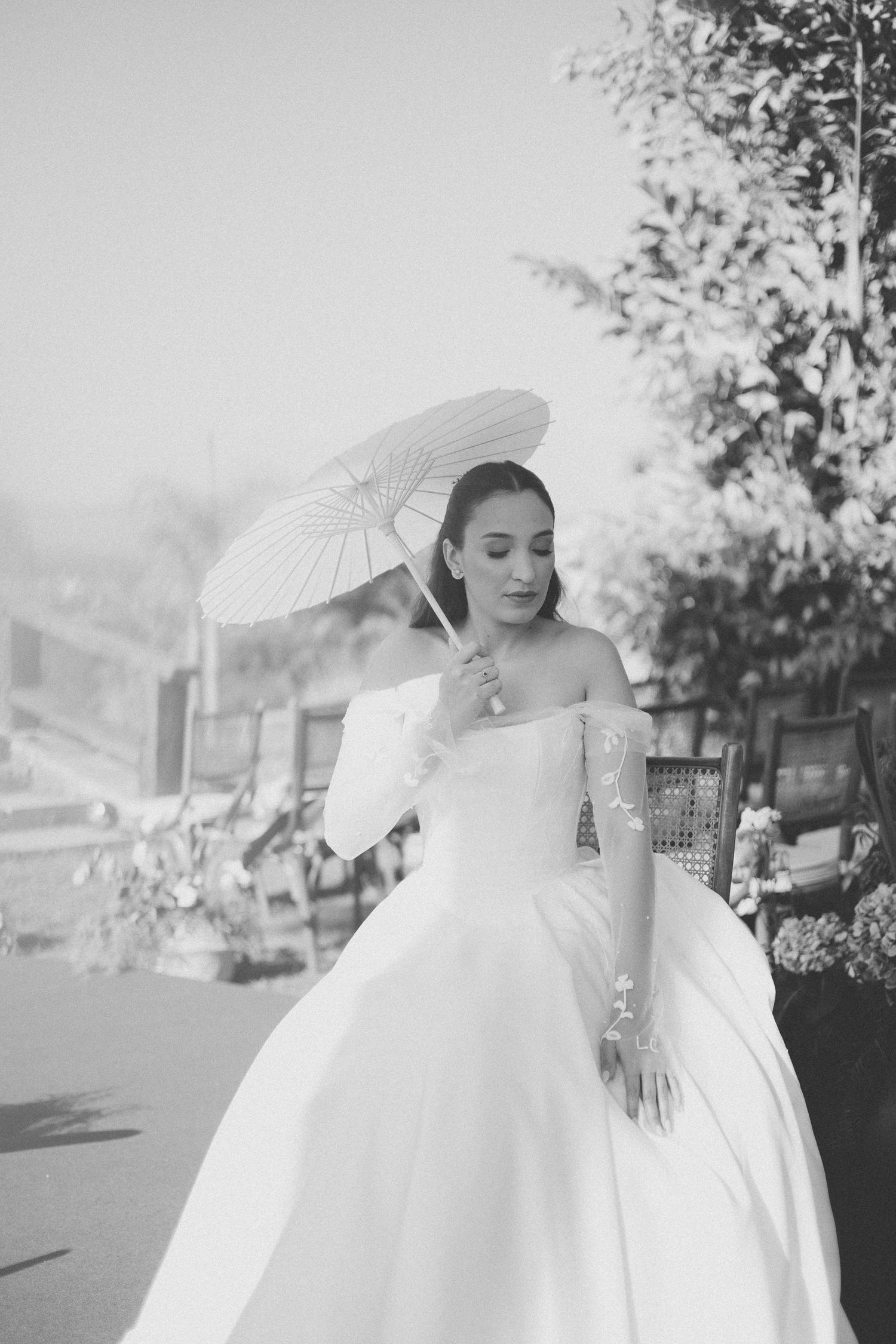 Black and white photo of a bride with a parasol in a flowing off-shoulder wedding dress on an outdoor terrace.