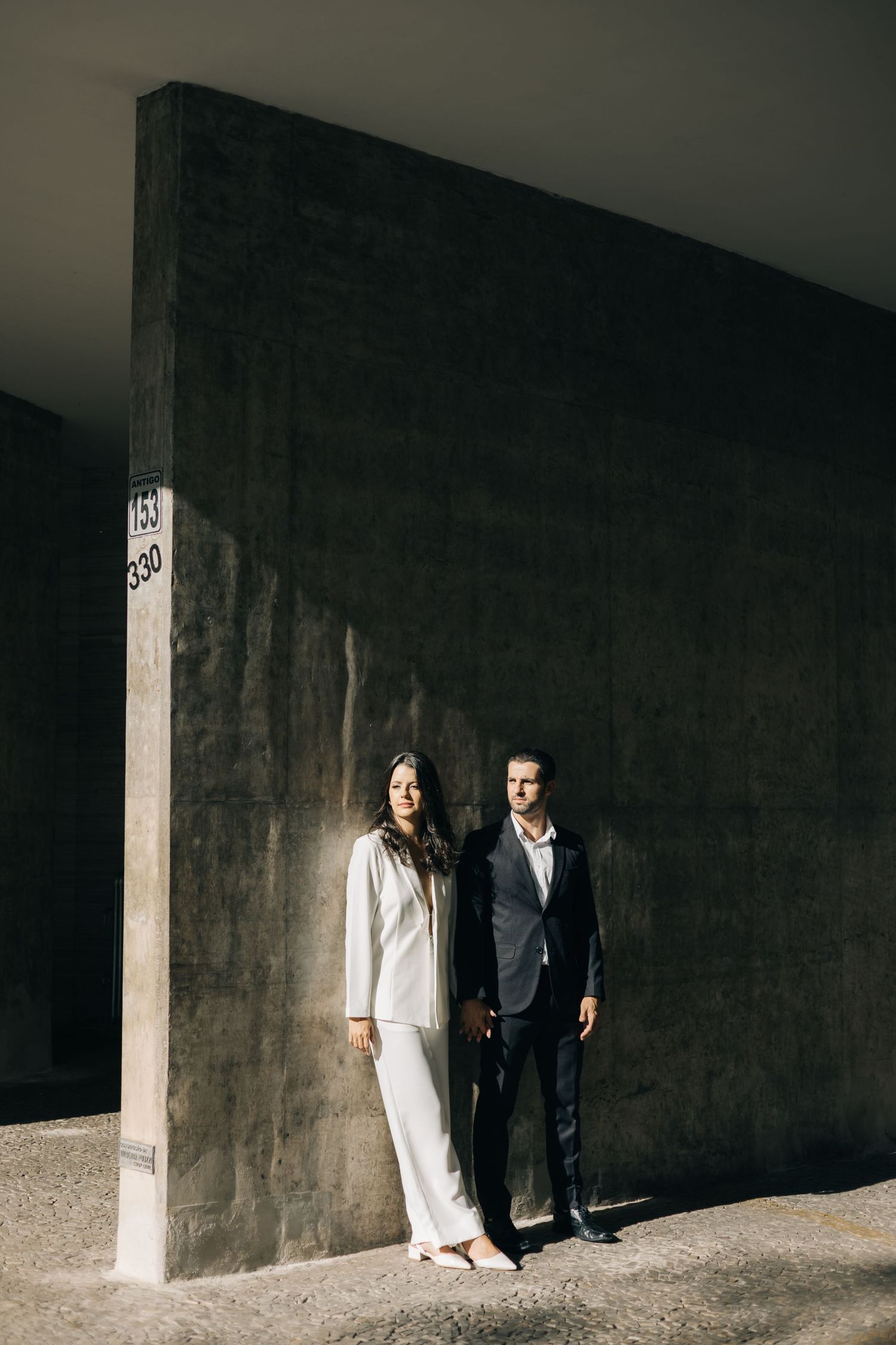 Two people in contrasting outfits of white and dark suits stand against a stark concrete wall in dramatic lighting.