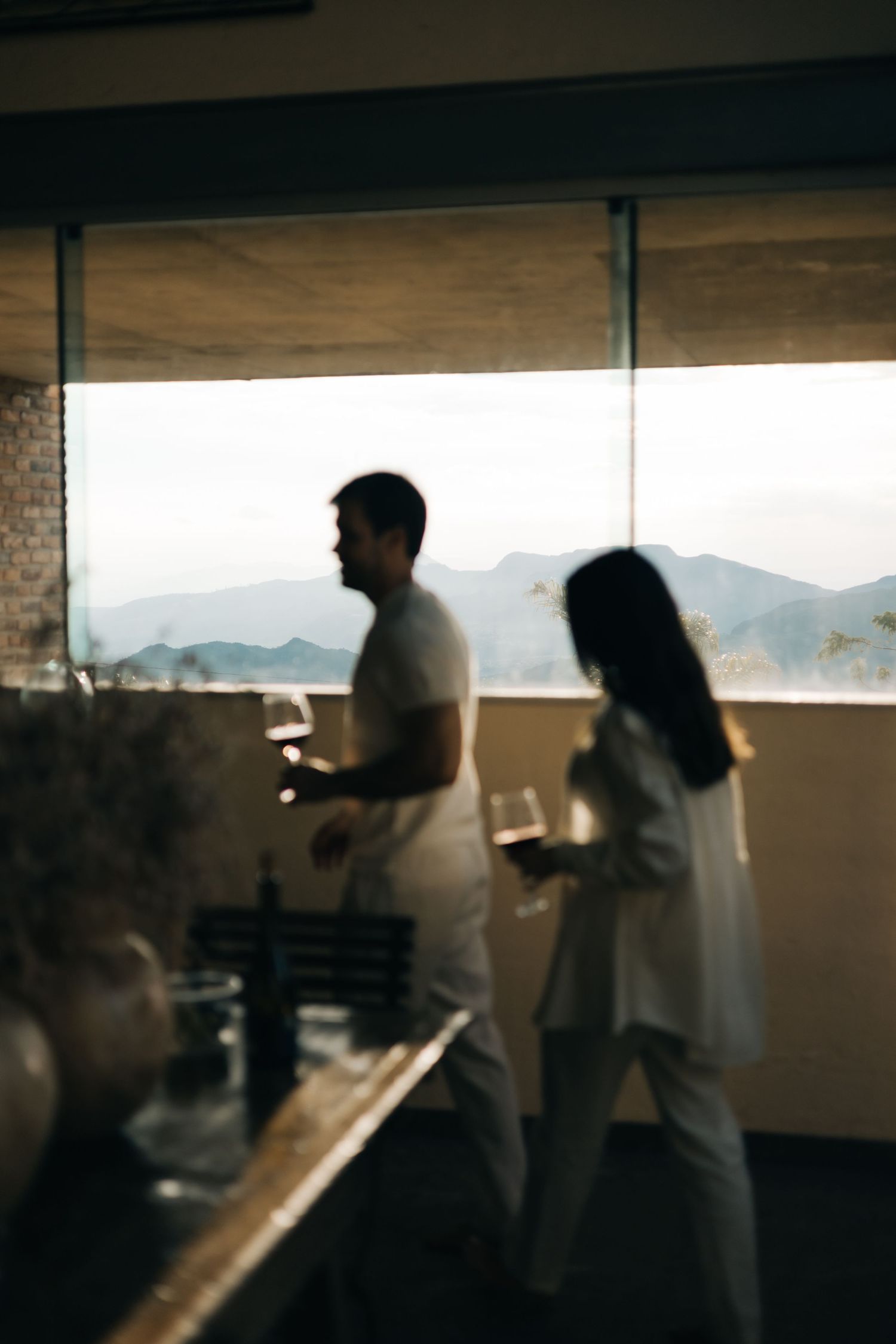 Two people enjoying drinks on a balcony with mountain views in the background on a hazy afternoon.
