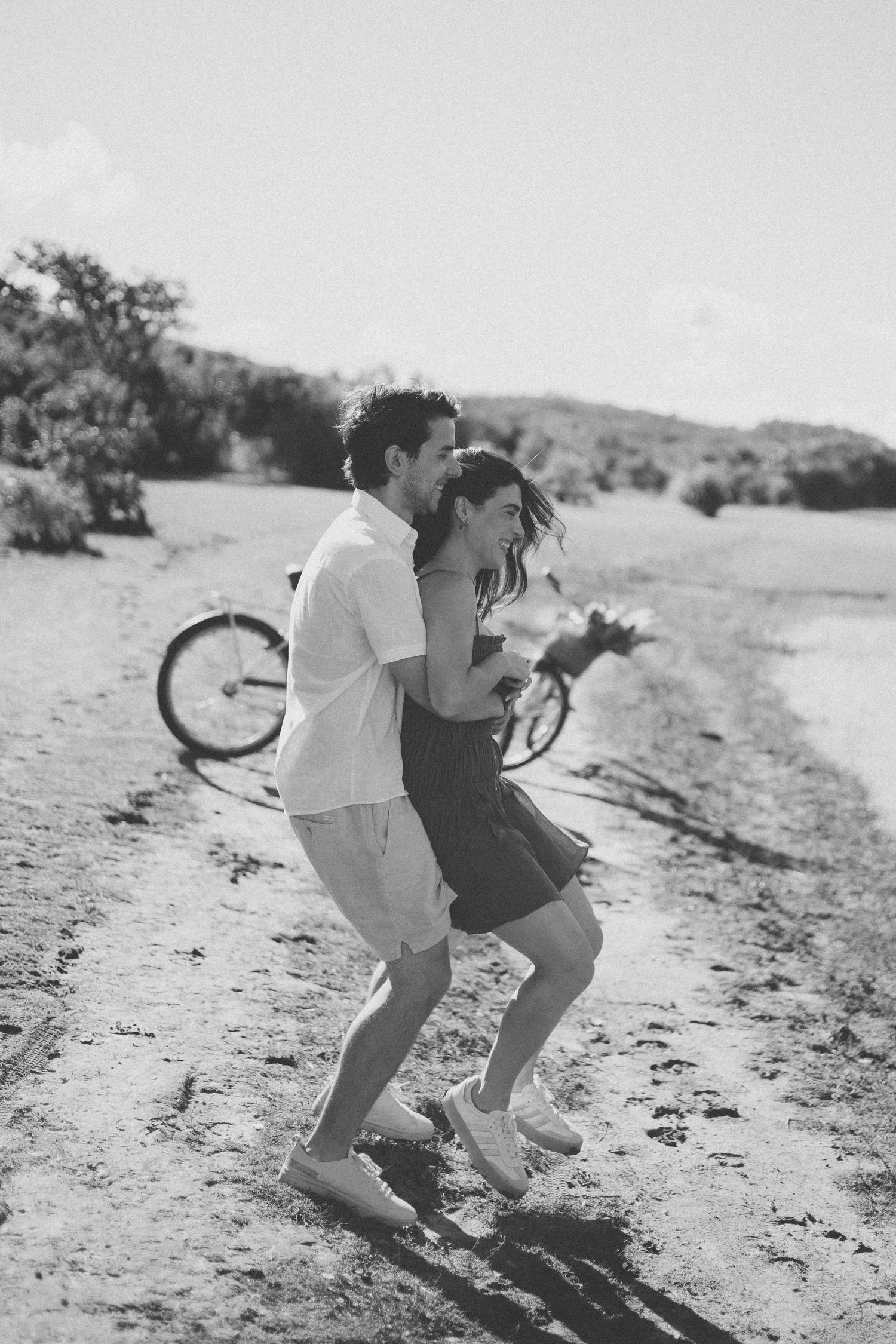 A couple dances together on a dirt path with a bicycle in the background on a sunny day.