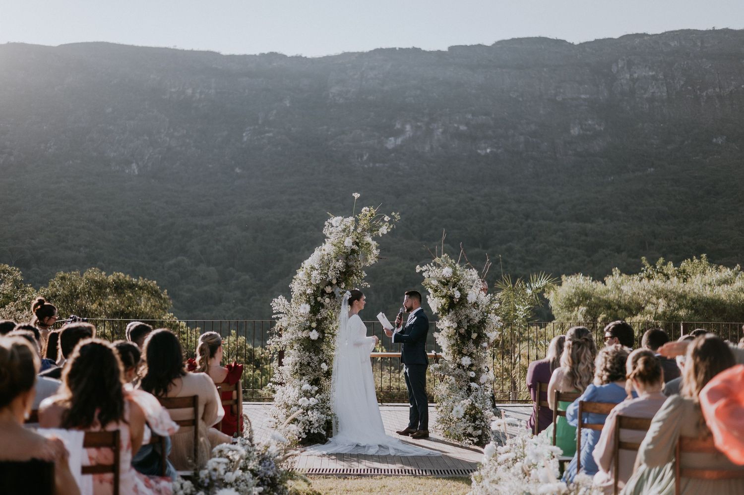 Wedding ceremony with floral arch overlooking mountain landscape as guests capture photos.