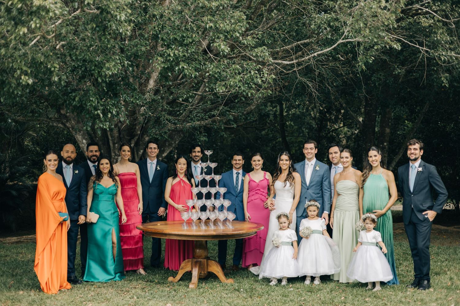 Wedding party members in colorful dresses pose with champagne tower outdoors.