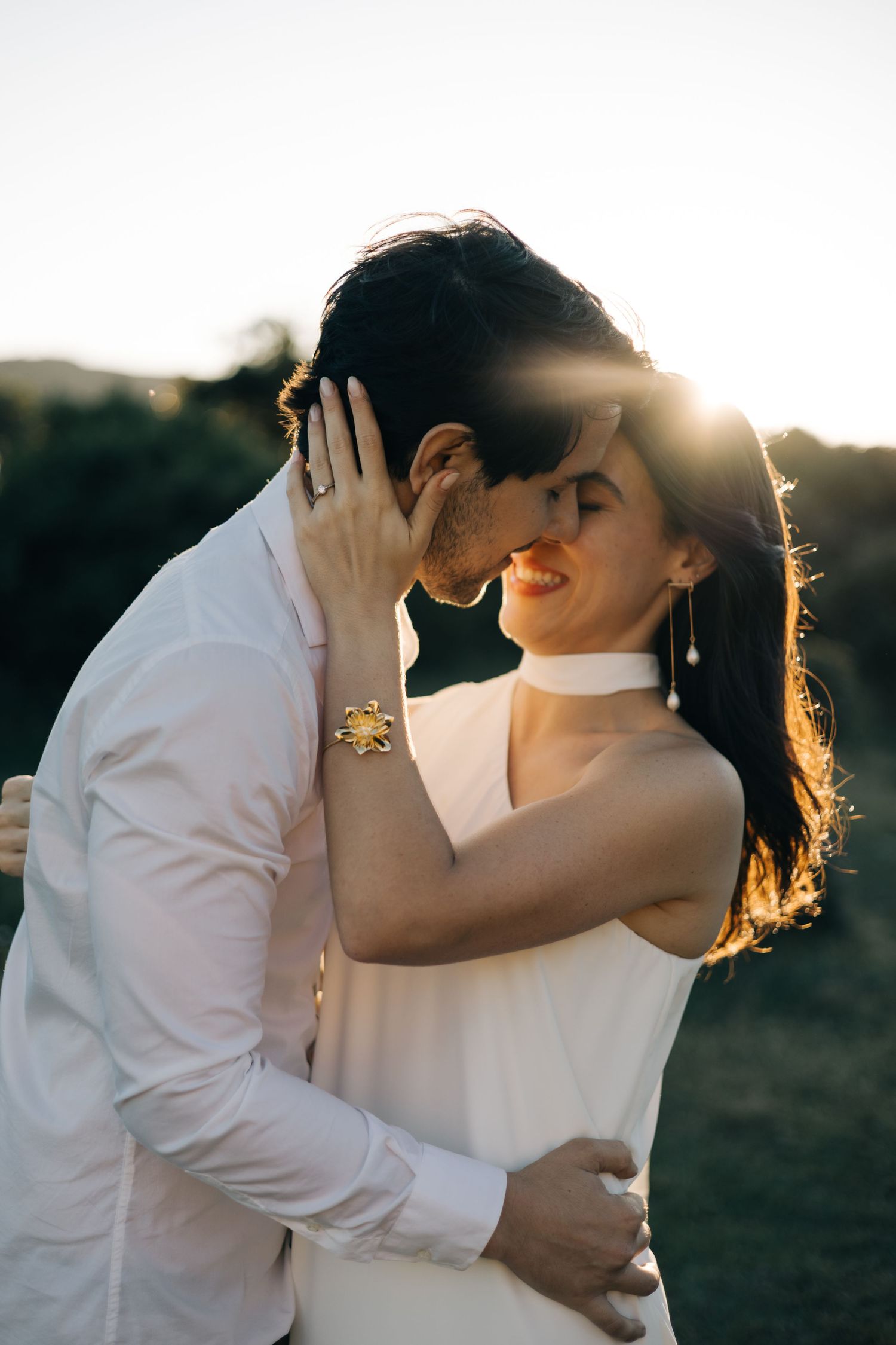 Romantic couple in white outfits embrace during golden sunset, with sun flare creating ethereal backlighting.