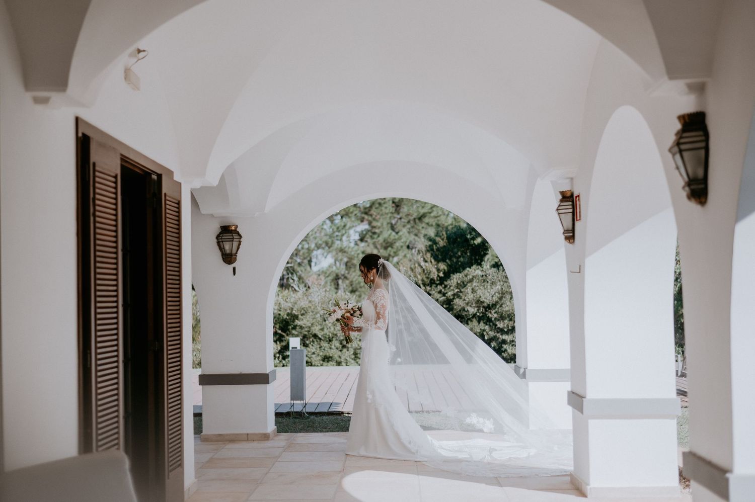 Elegant bridal portrait under white archway with flowing veil in natural light.