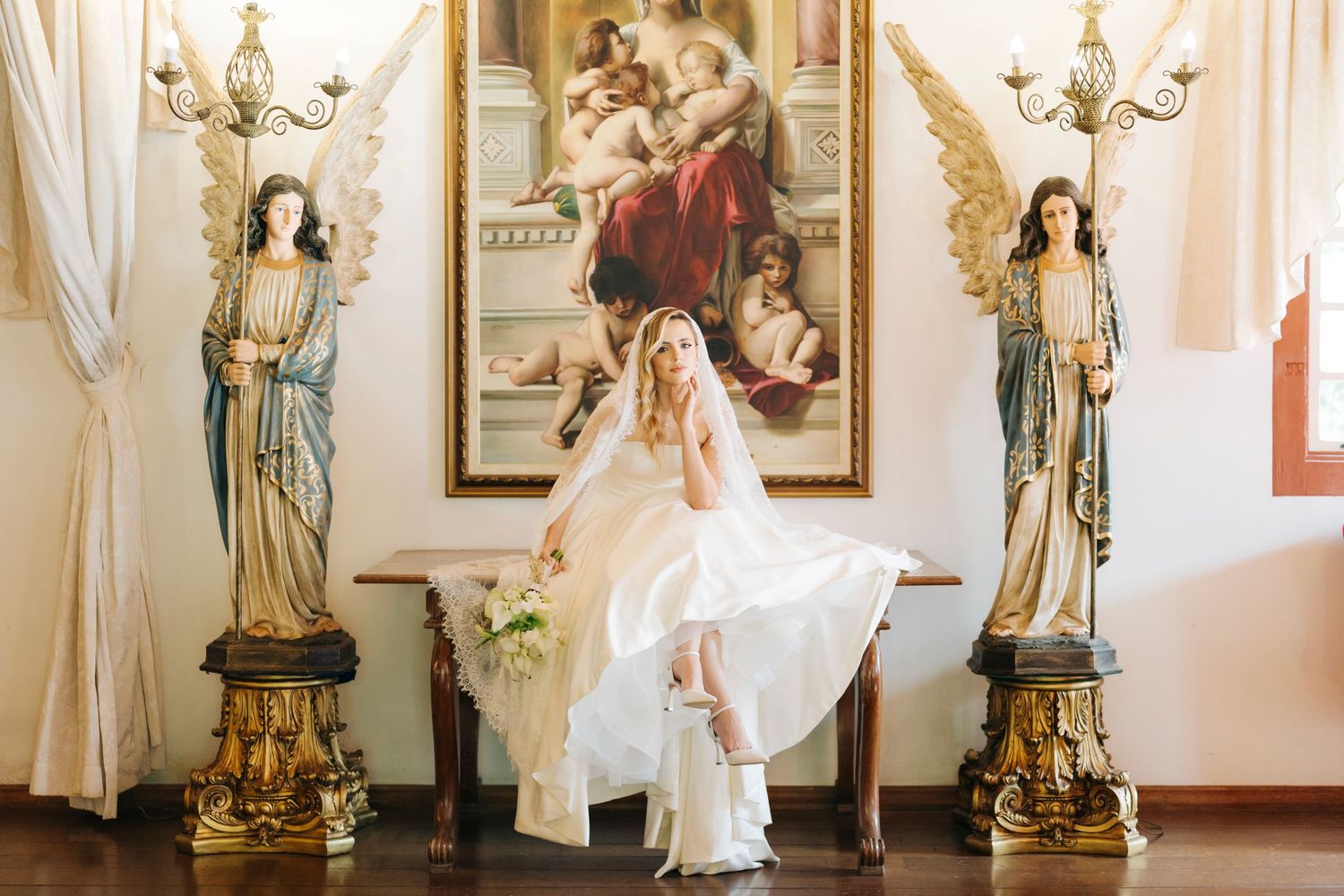 Bride in white gown poses between angel statues with religious painting backdrop in elegant venue.