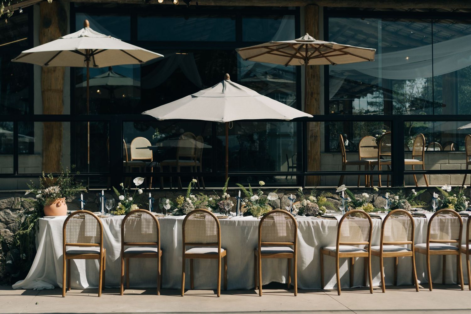 Elegant outdoor dining setup with long table under white market umbrellas against modern glass building backdrop.