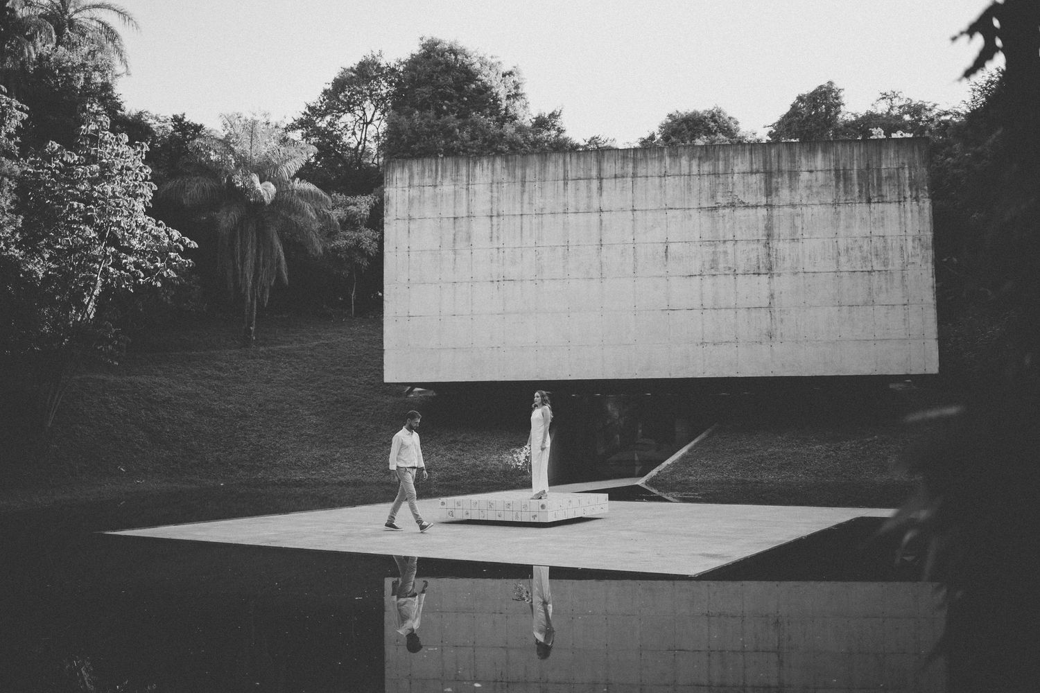 Black and white minimalist scene of two people standing on a reflective surface in front of a blank white wall.