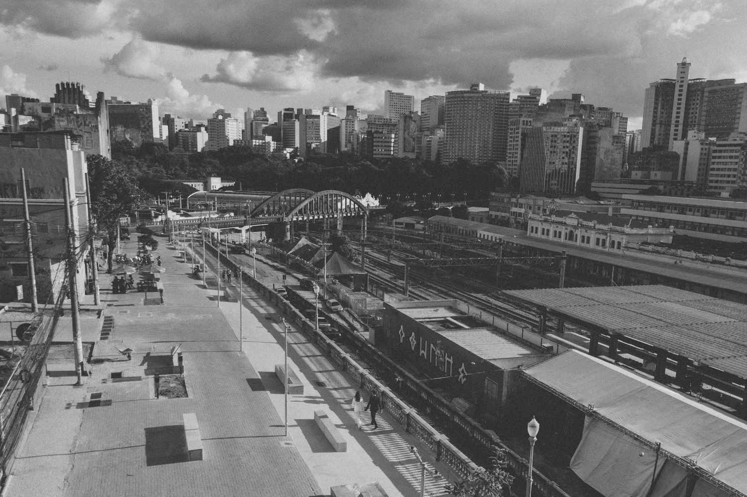 Black and white aerial view of urban train tracks and platforms with city buildings in the background under cloudy skies.