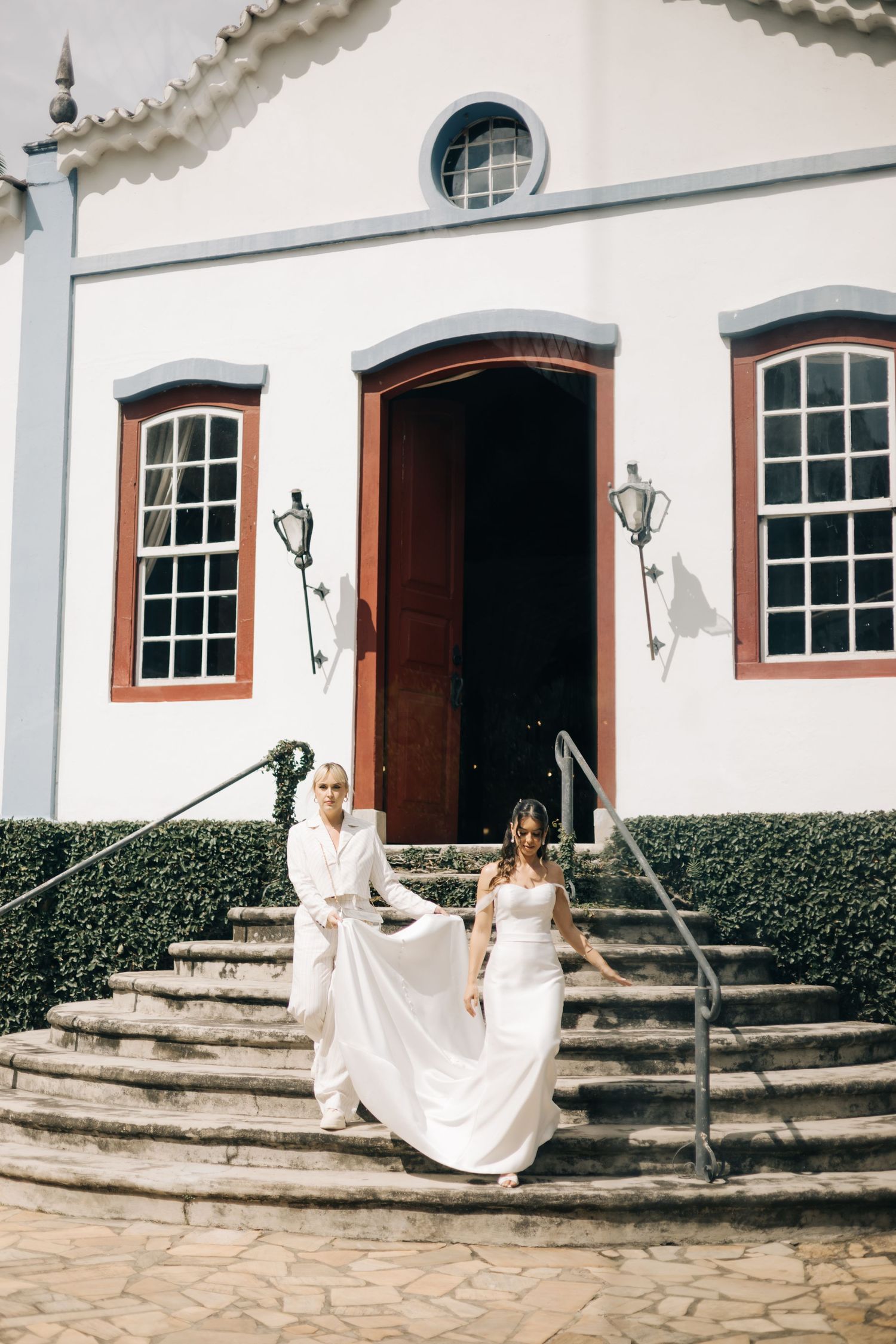 Bride descending white church steps while attendant helps with long train of dress.