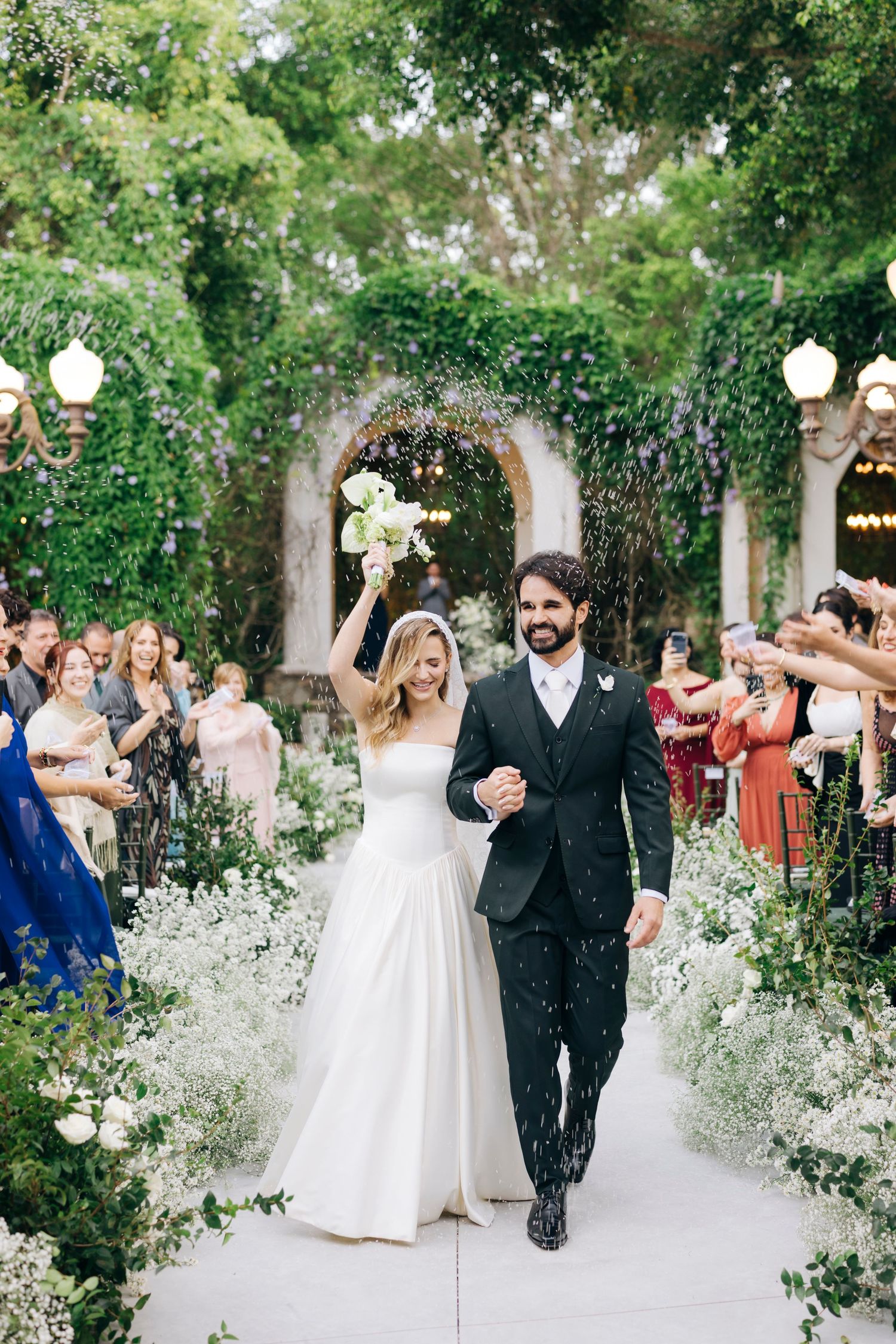 Bride and groom walk down flower-lined aisle after wedding ceremony, surrounded by cheering guests in lush garden venue.