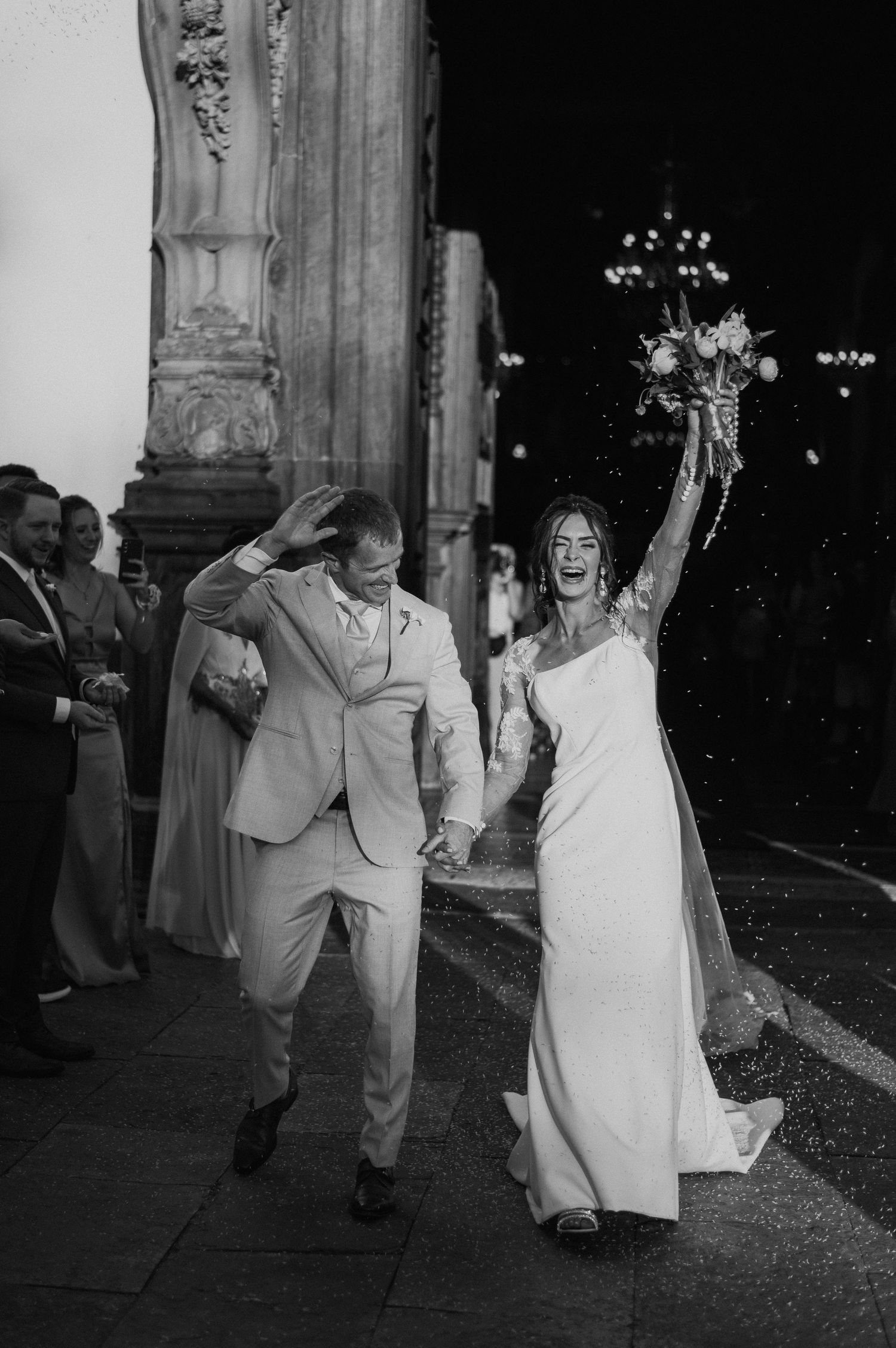 Joyful newlyweds exit church with raised bouquet in black and white photograph.