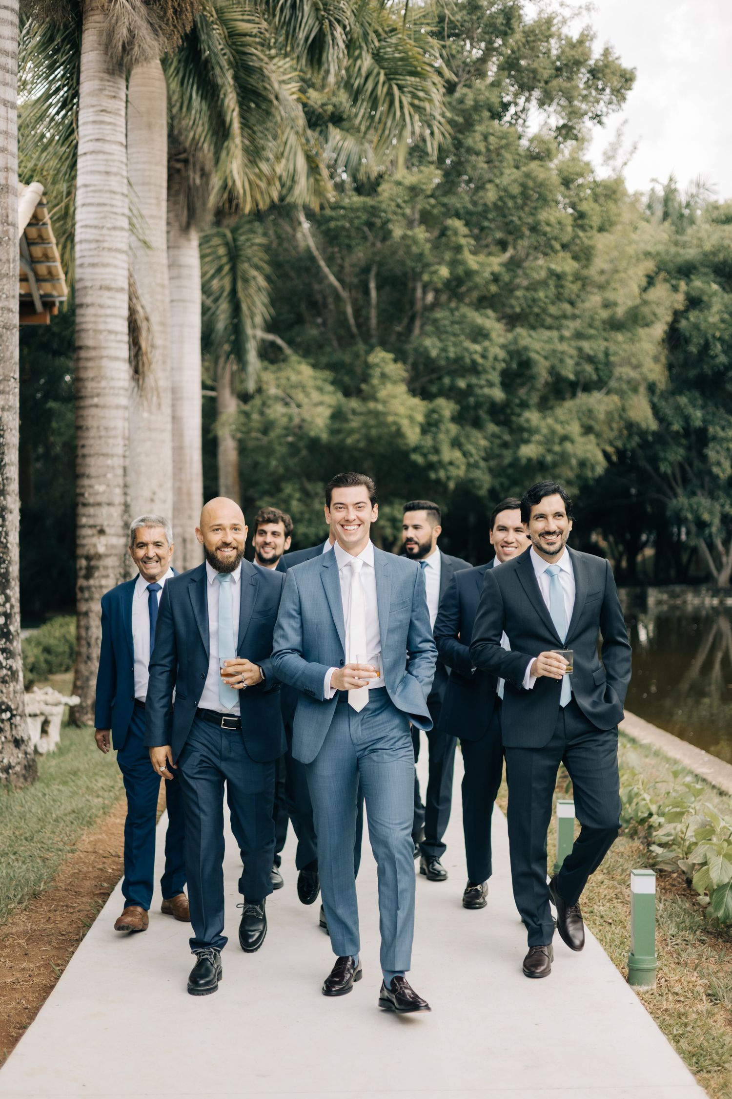Groomsmen in navy suits walking down a palm tree-lined pathway led by the groom in light blue.