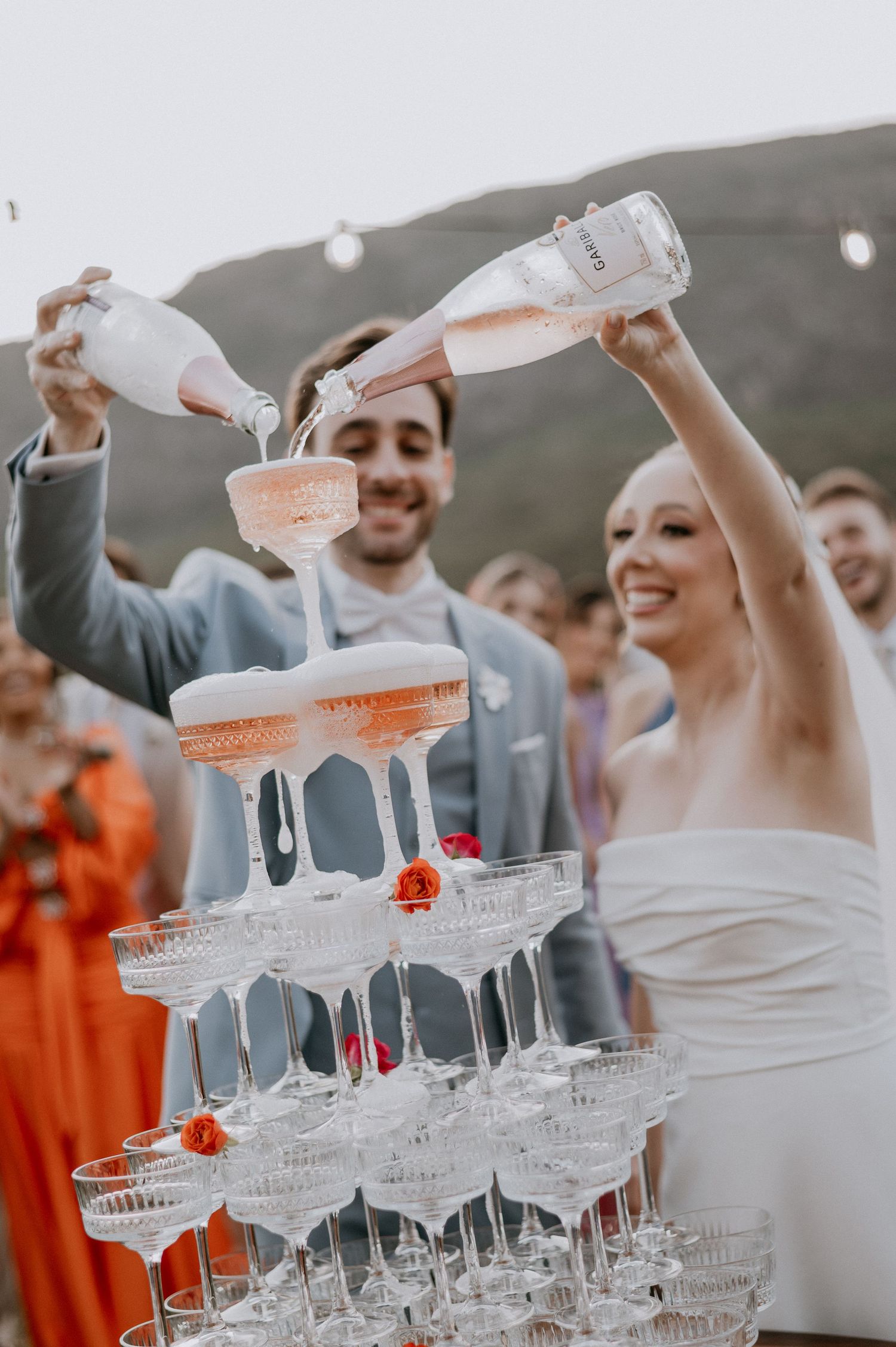 Champagne being poured into tower of crystal glasses at outdoor wedding celebration.