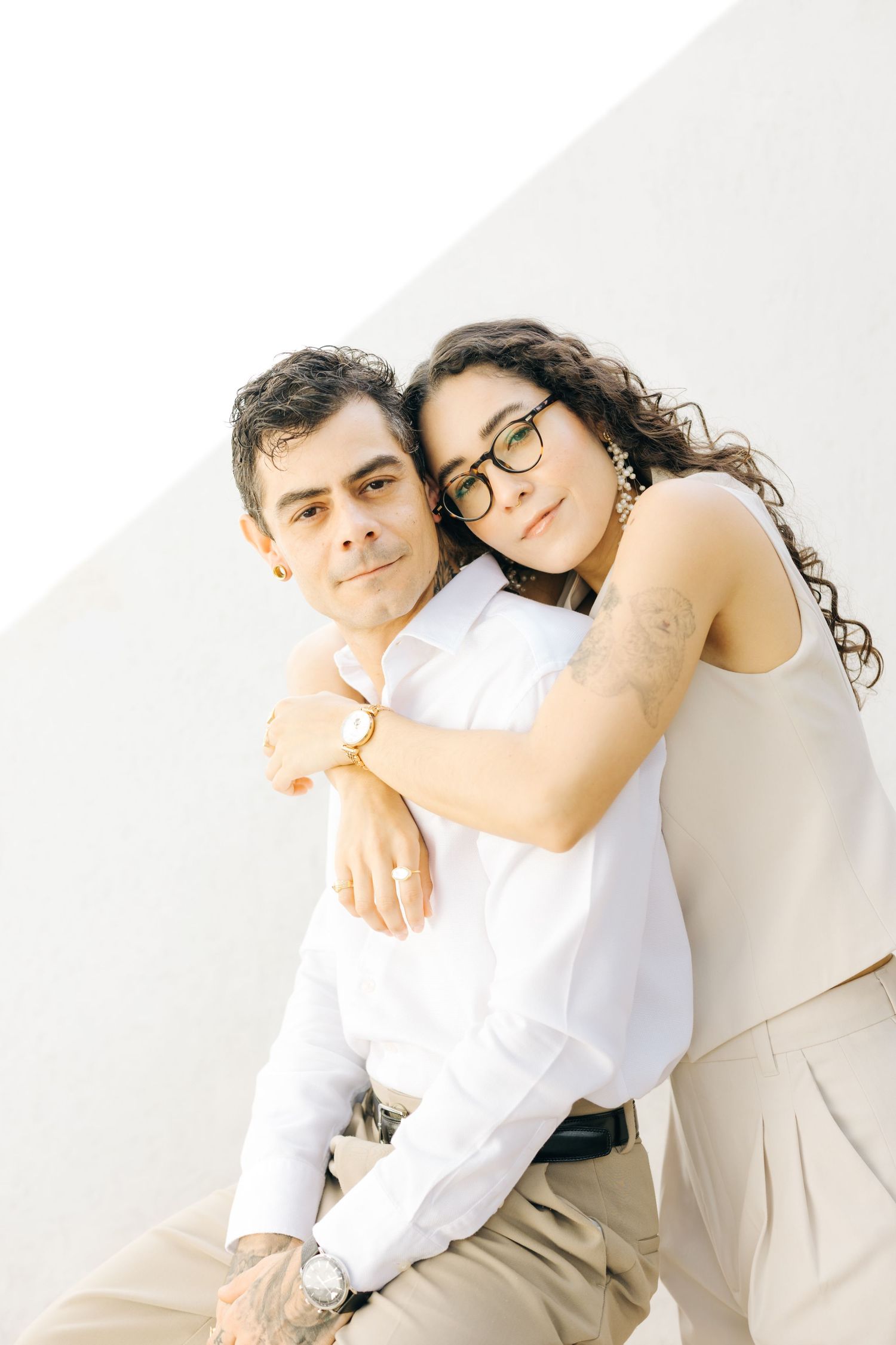 A couple embracing warmly, woman with glasses hugging man in white shirt, posing against a light background.