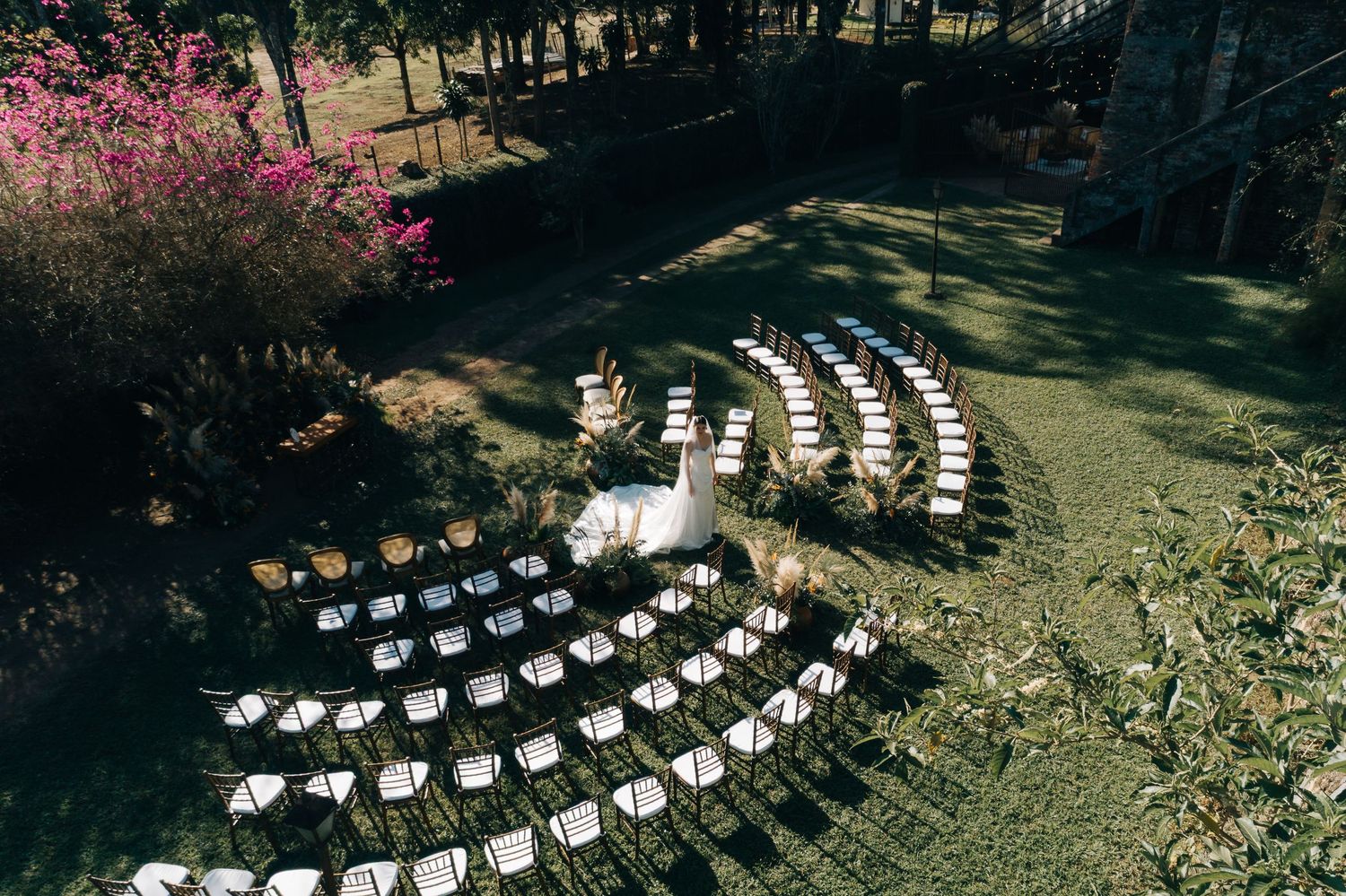White wedding chairs arranged in curved rows on a lawn with blooming pink trees and forest backdrop for outdoor ceremony.