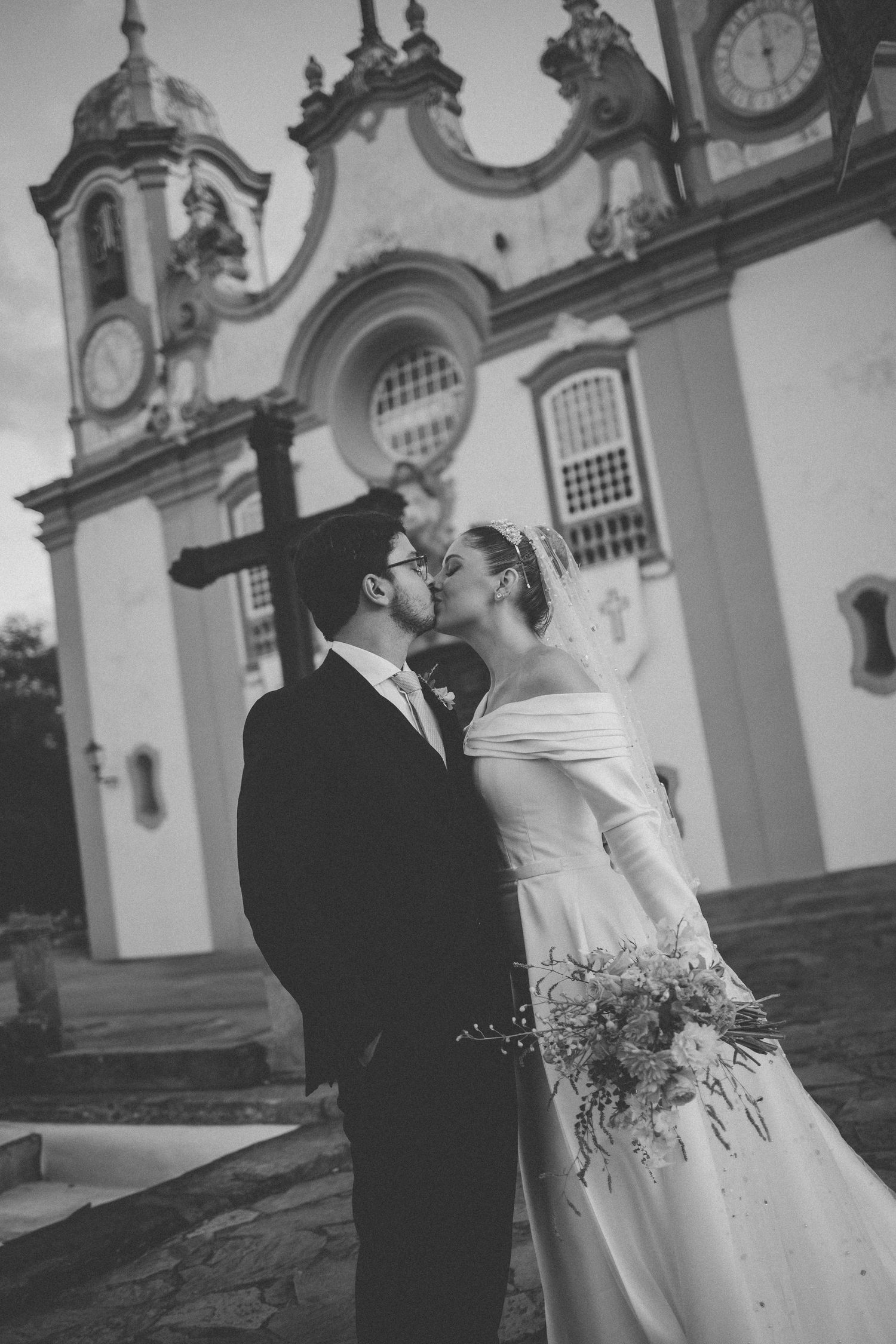 Black and white photo of a bride and groom kissing in front of a historic baroque church, bride holding a bouquet.