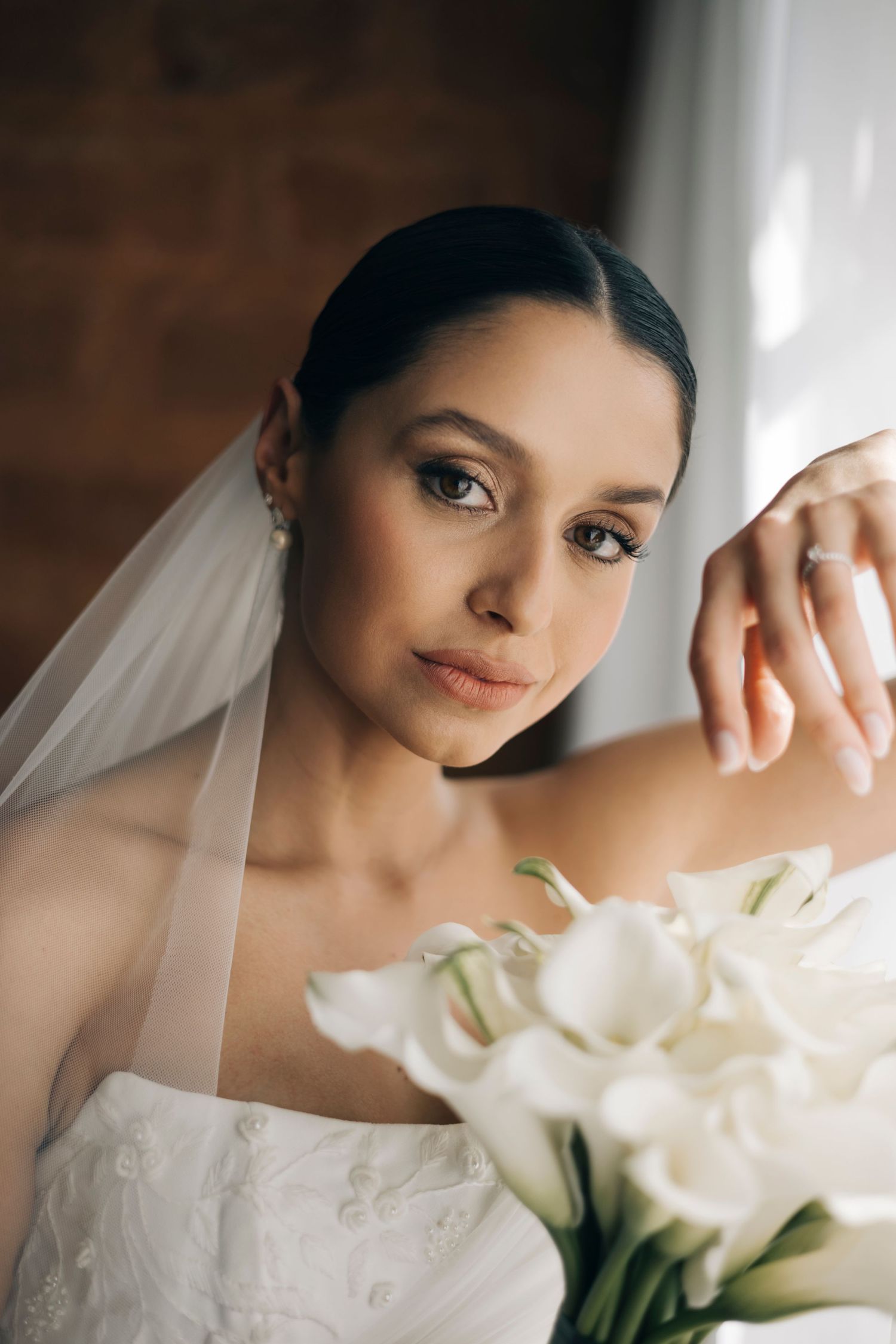 Elegant bride in white strapless gown and veil holding calla lily bouquet, wearing diamond ring, posing by window.