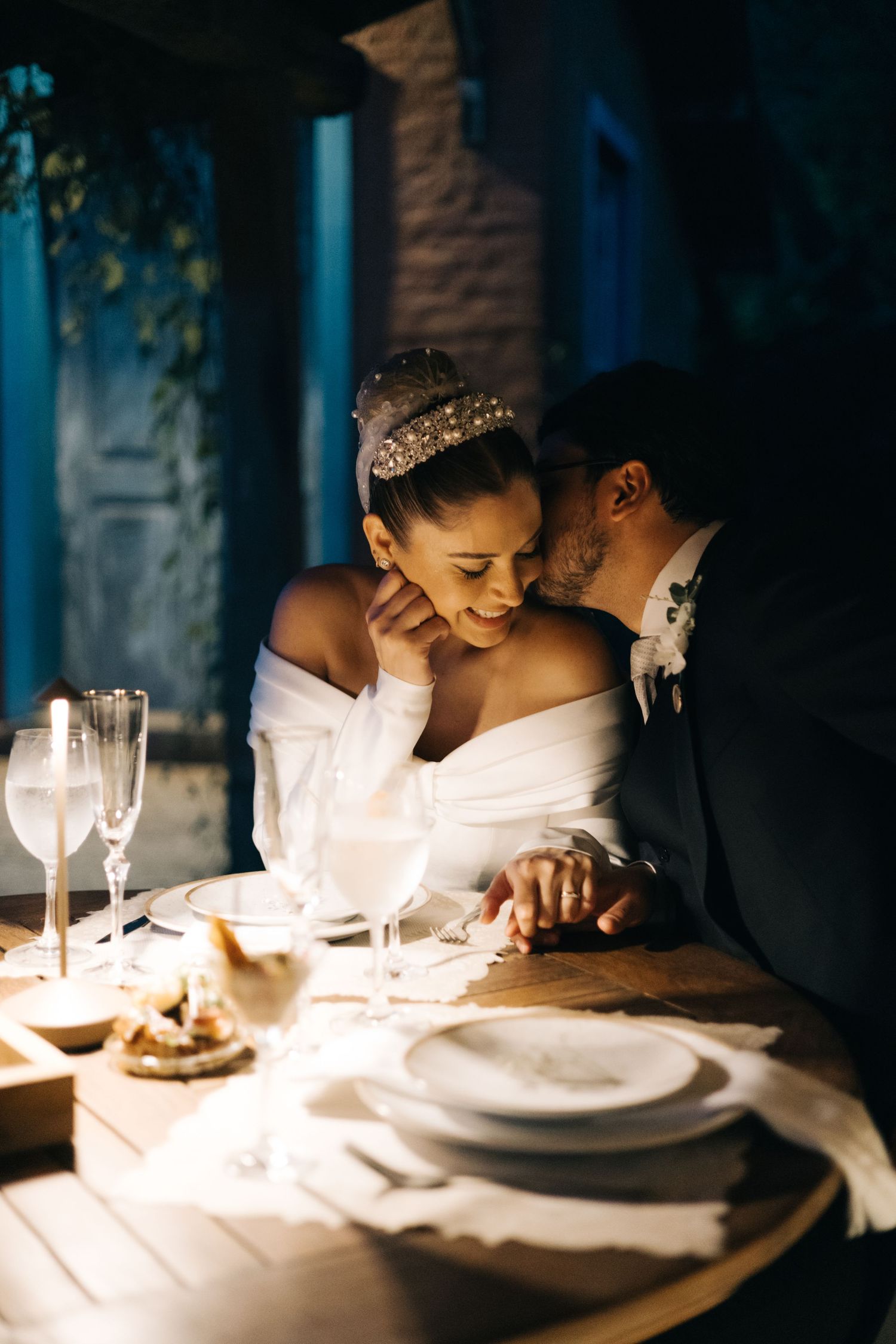 Bride and groom share an intimate moment at candlelit wedding reception table, groom kissing bride's cheek.