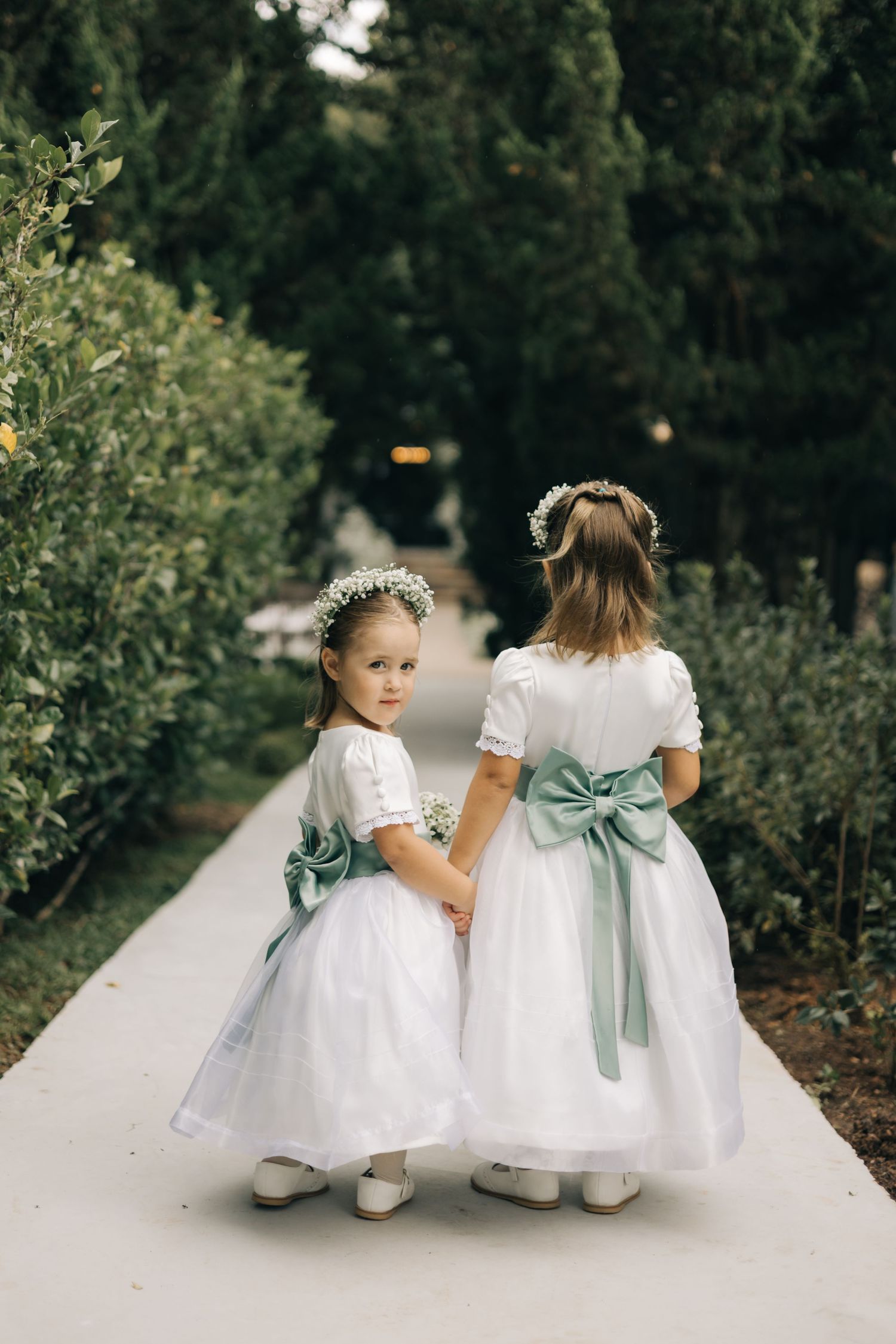 Two flower girls in white dresses with mint green sashes hold hands on a garden pathway.