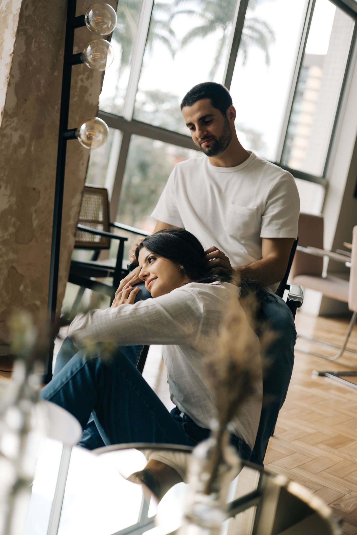 Two individuals in casual clothing share a tender moment near large windows as soft light streams through from outside.