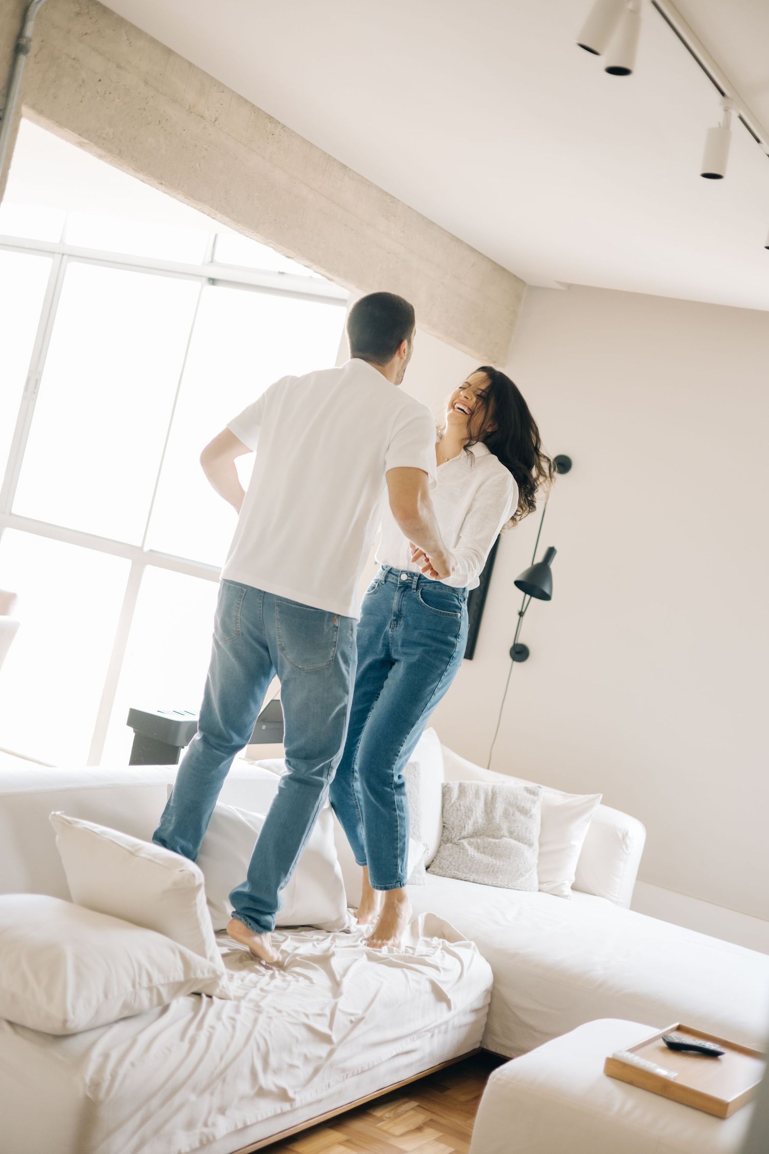Two people in casual outfits having fun jumping on a white sofa in a bright minimalist living room.