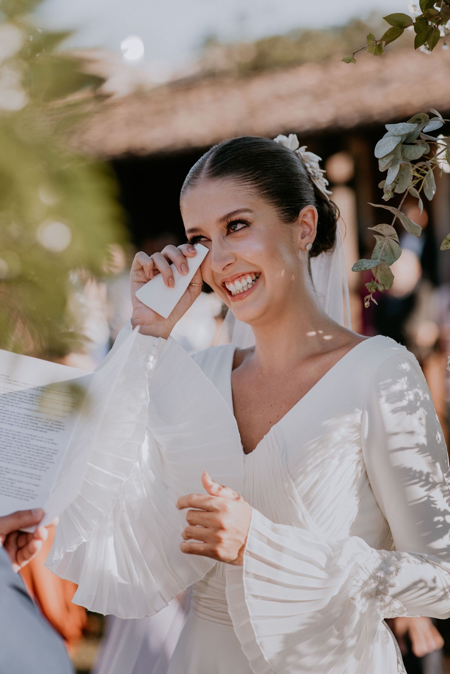 Emotional bride in white dress wiping tears during outdoor wedding ceremony.