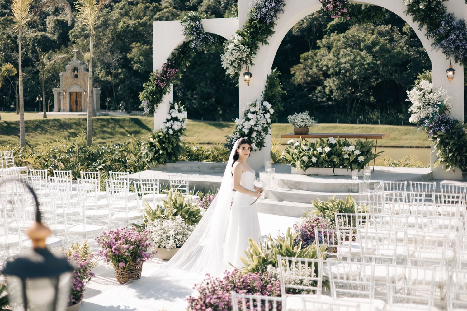 Elegant outdoor wedding ceremony setup with white chairs, floral decorations, and a beautiful arched pergola in a garden setting.