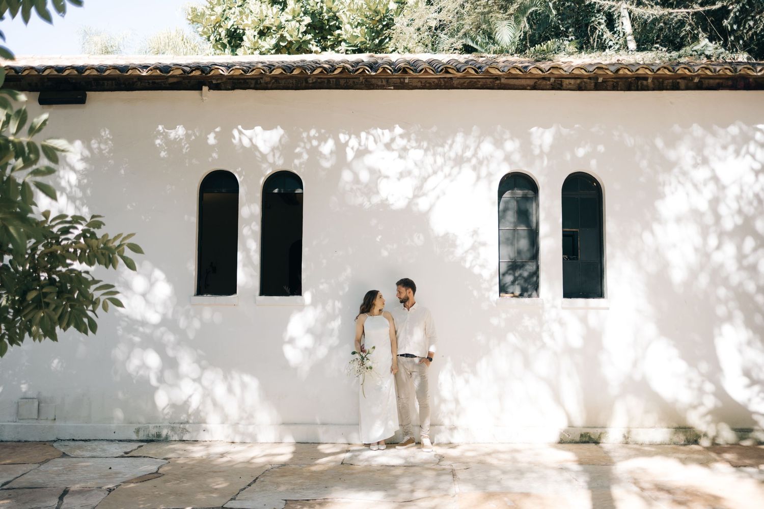 A couple stands together against a white building with arched windows casting dappled shadows on the wall.