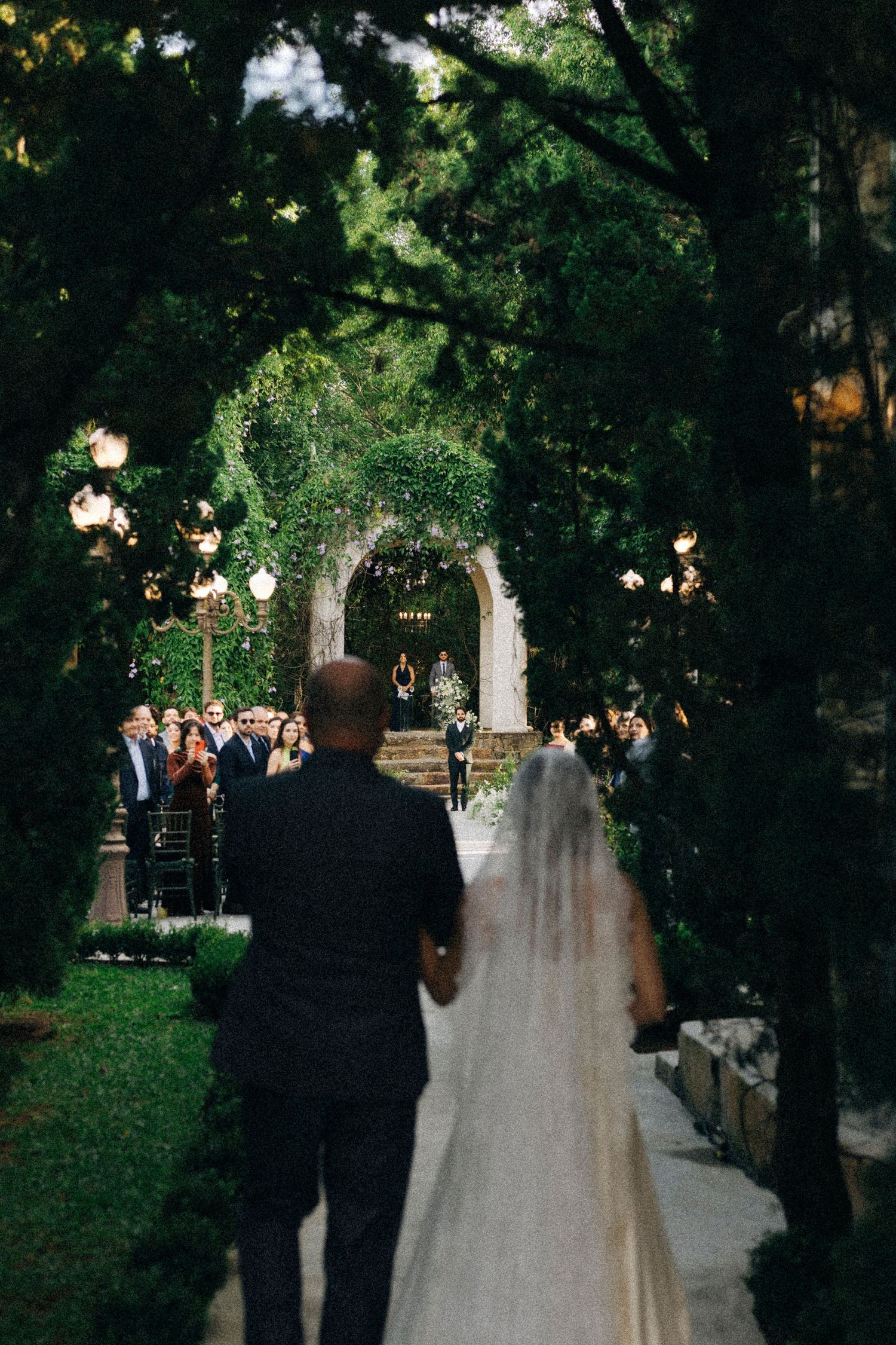 Bride in white gown and veil walks down outdoor aisle with groom toward ivy-covered arch at elegant garden wedding ceremony.