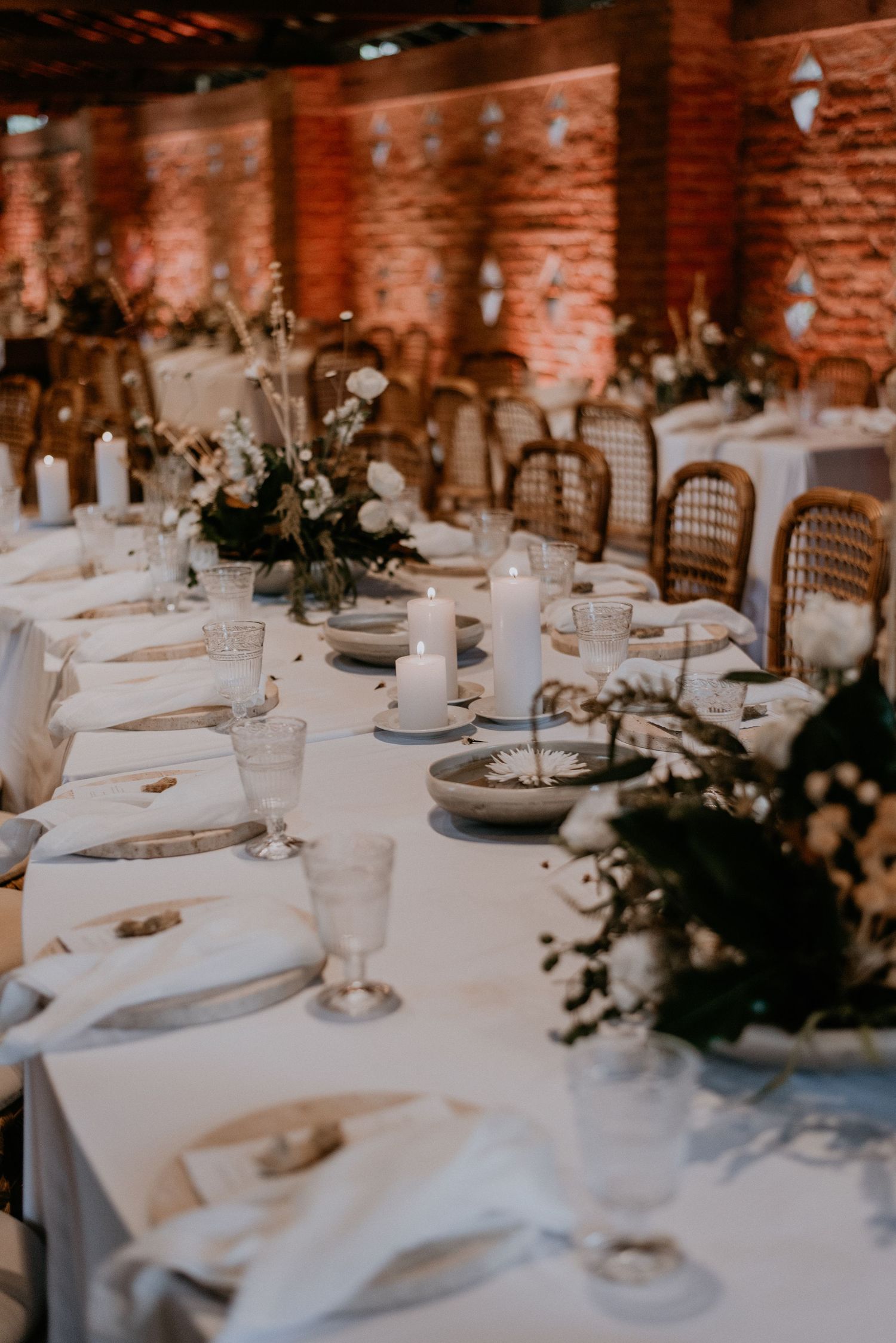 Elegant wedding reception table setting with white linens and rattan chairs in rustic brick venue.