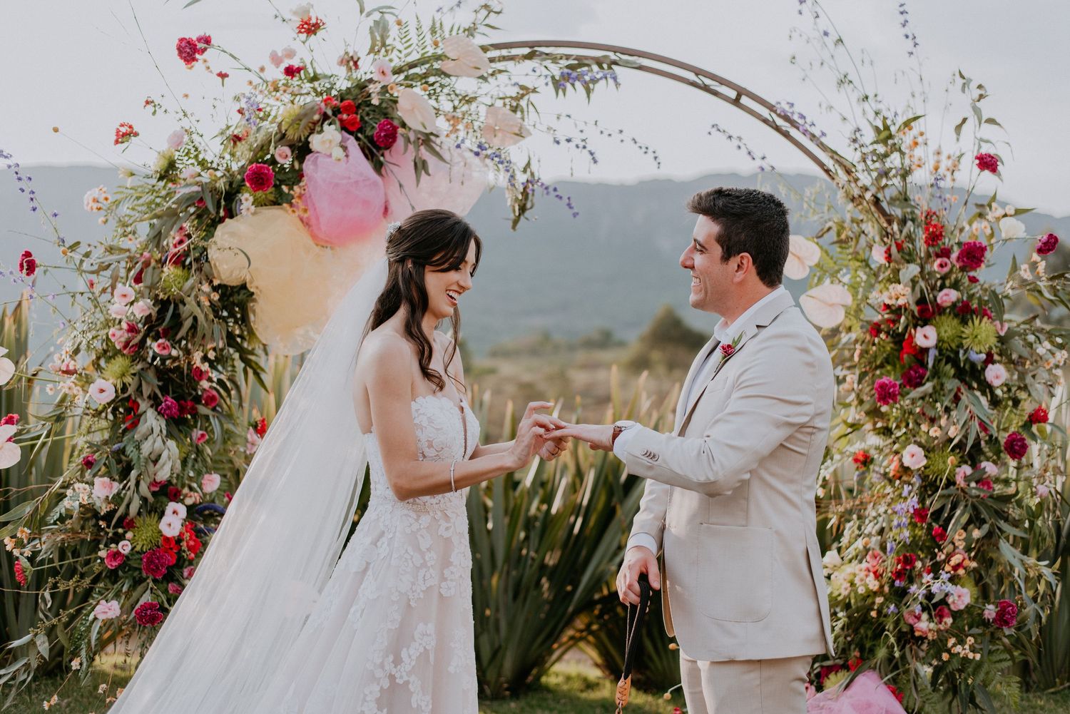 Couple exchanging vows under circular floral arch with pink accents and mountain scenery.