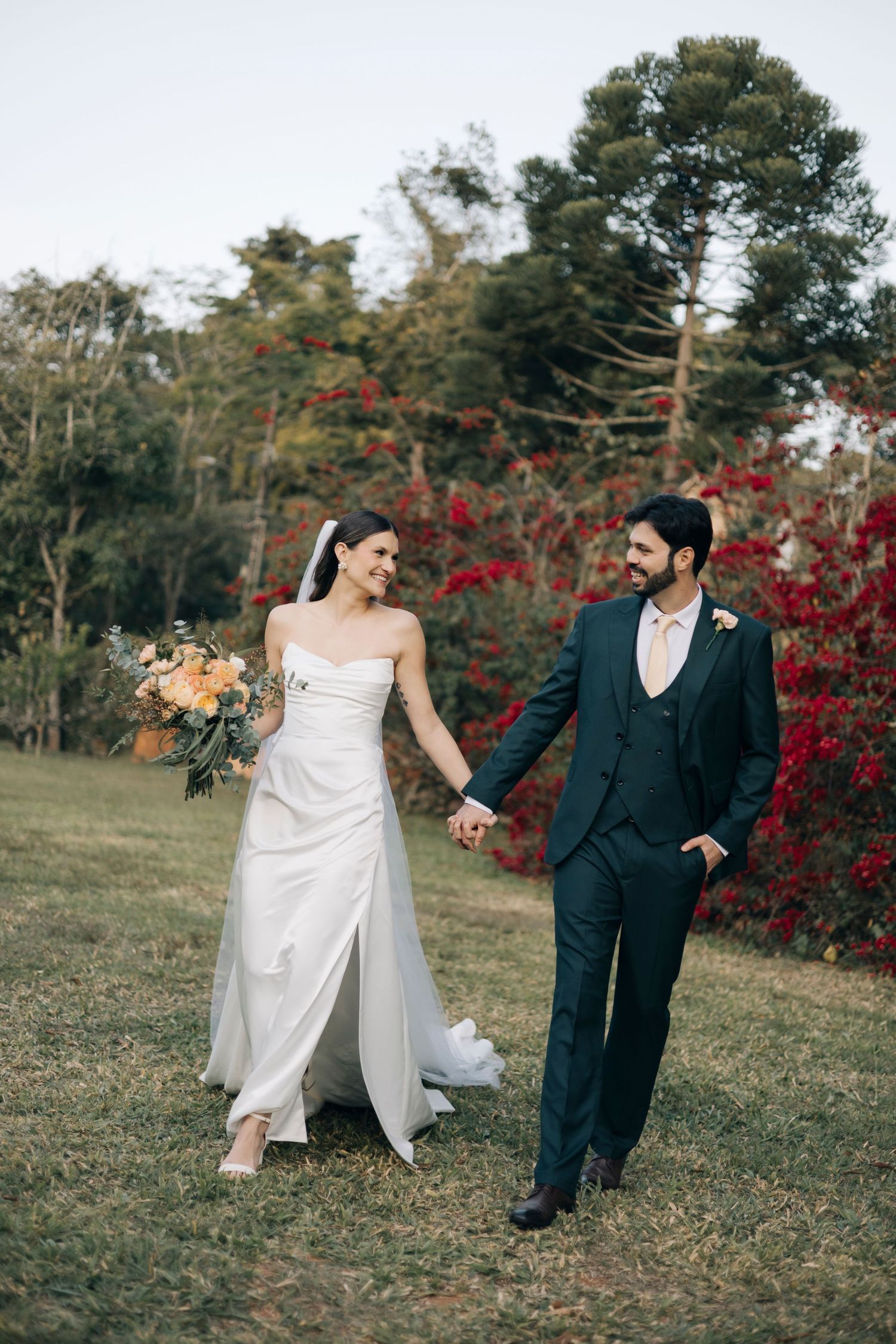 A couple in wedding attire walks hand in hand through a garden with vibrant red autumn foliage in the background.