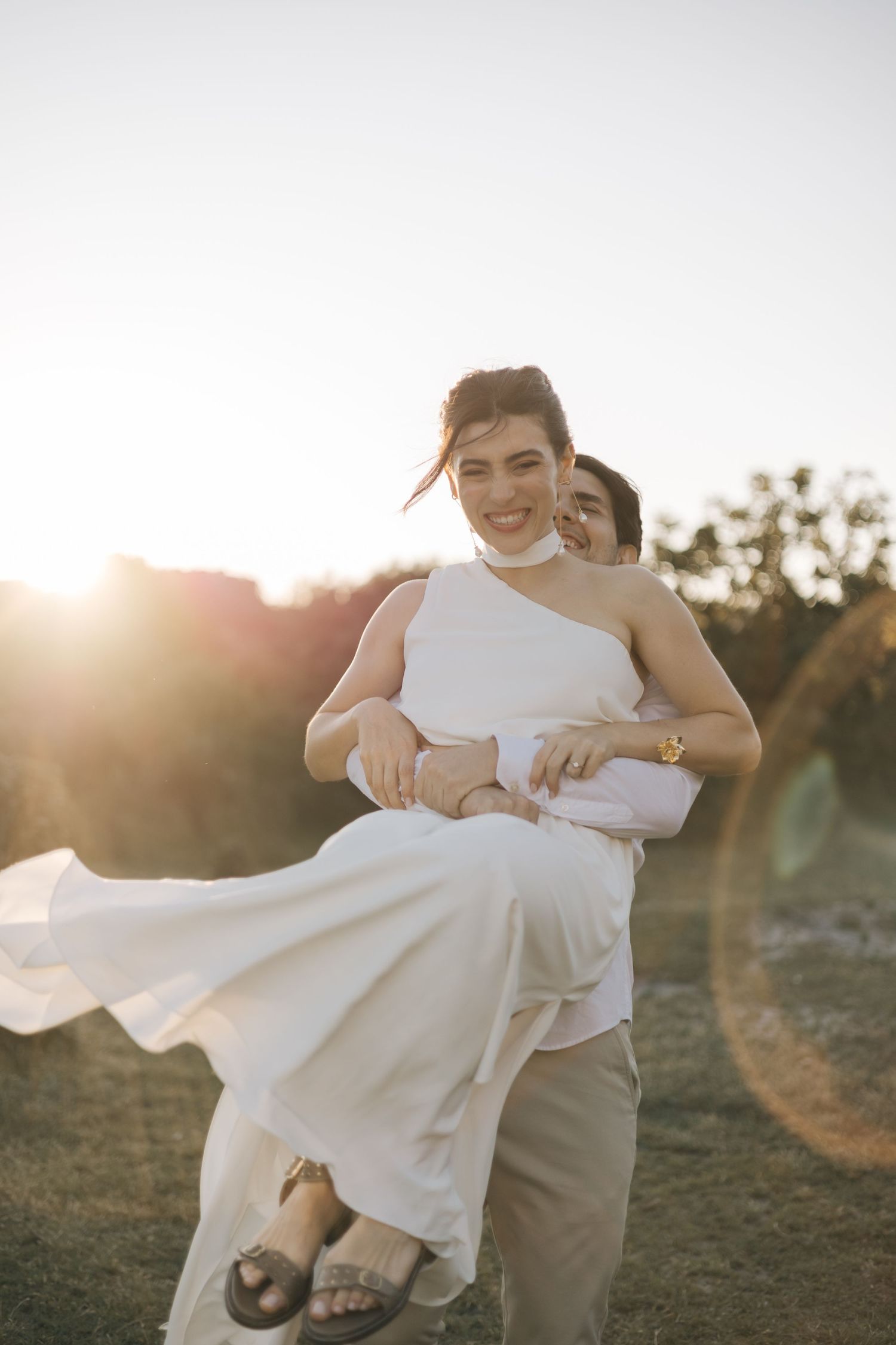 Two people embrace outdoors in golden sunlight with one wearing a flowing white wedding gown.