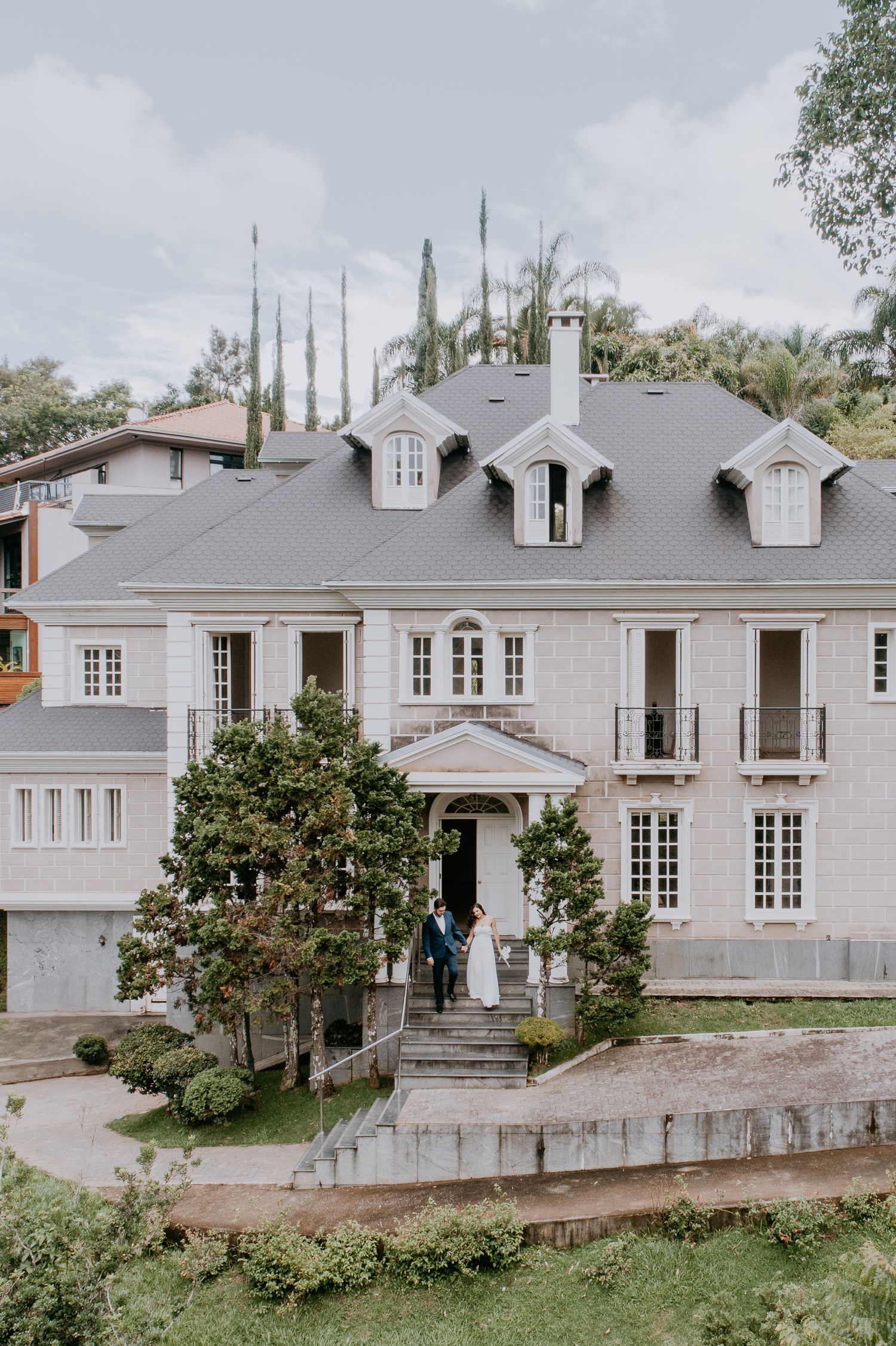 A grand colonial-style house with gray walls, white trim, and dormer windows sits on a landscaped hillside property.