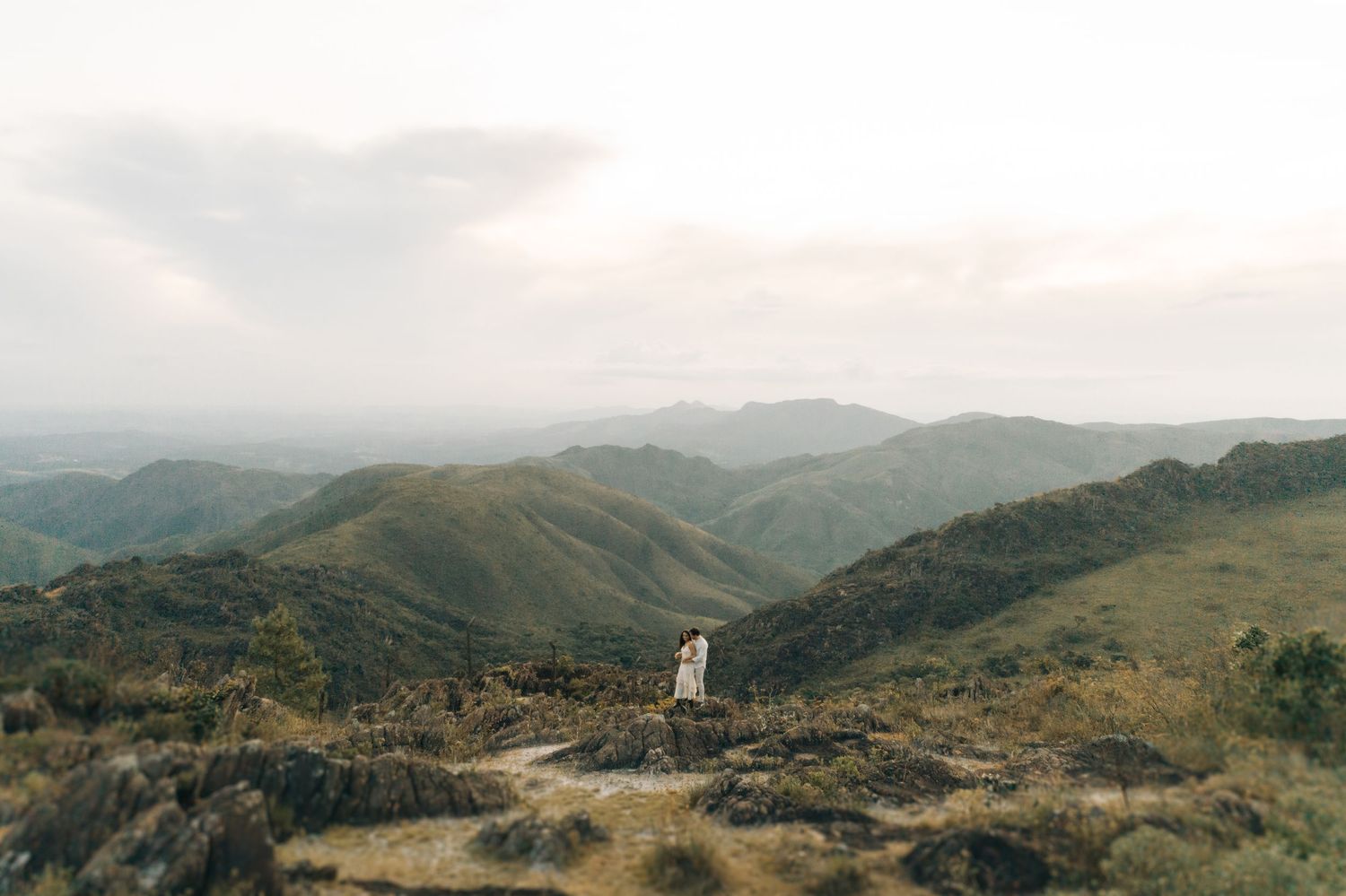 A small white structure stands alone on a rugged hilltop overlooking rolling mountains in the distance during sunset.