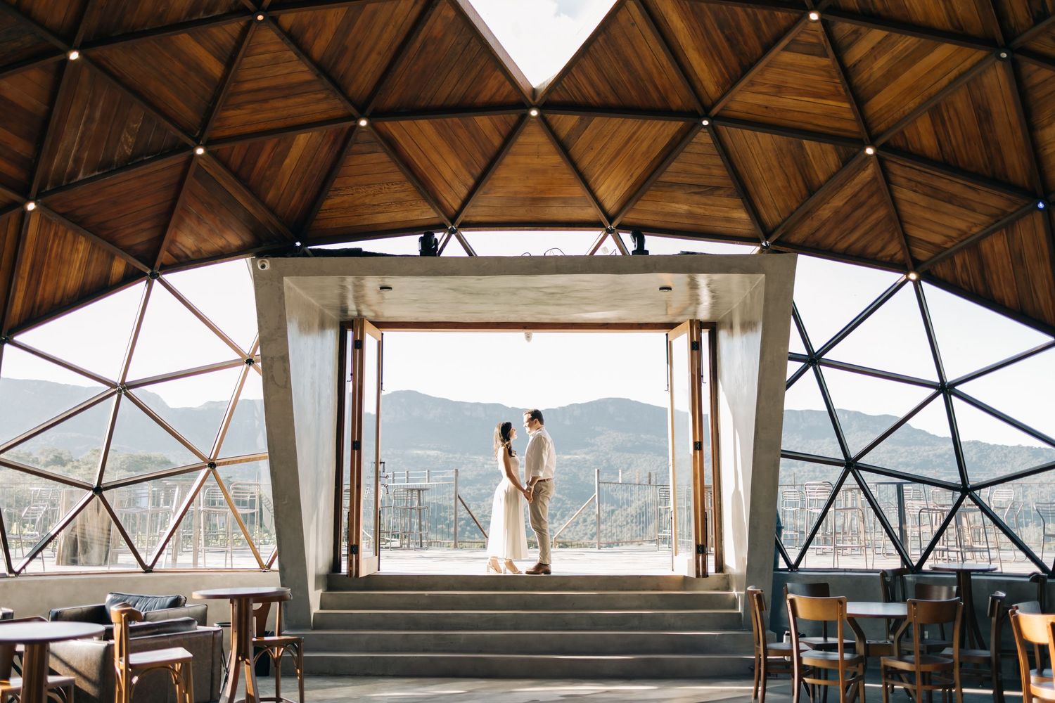 Two silhouettes stand in a geometric dome structure overlooking mountain vista through large glass windows.