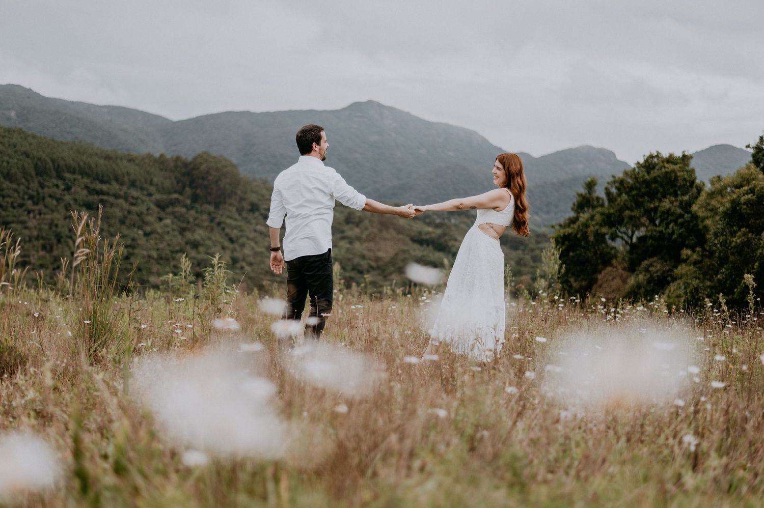 A couple stands together holding hands in a mountainous meadow with wildflowers and mountains in the background.