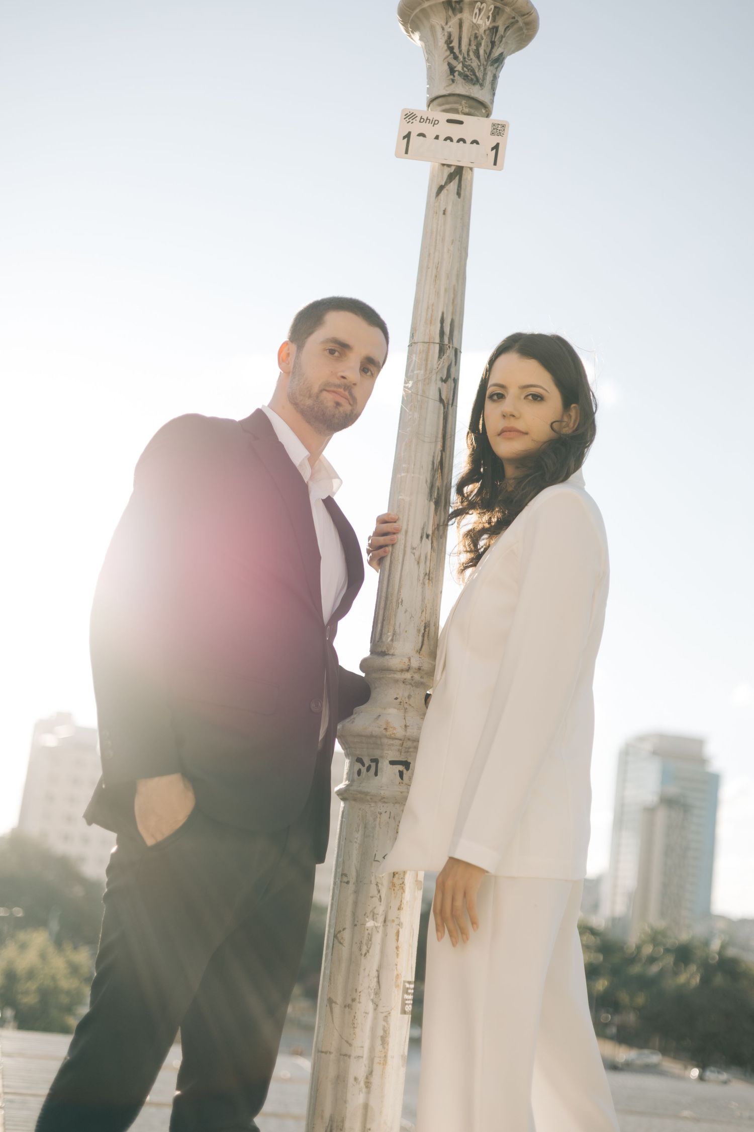 Elegant couple in formal attire poses beside vintage light post in urban setting with sunlit cityscape in background.
