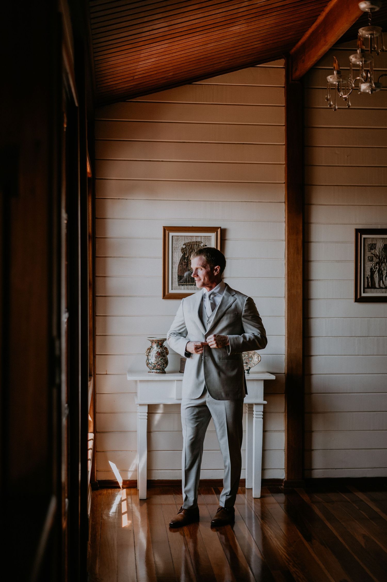 Groom in light gray suit stands by white table in rustic wood-paneled room.