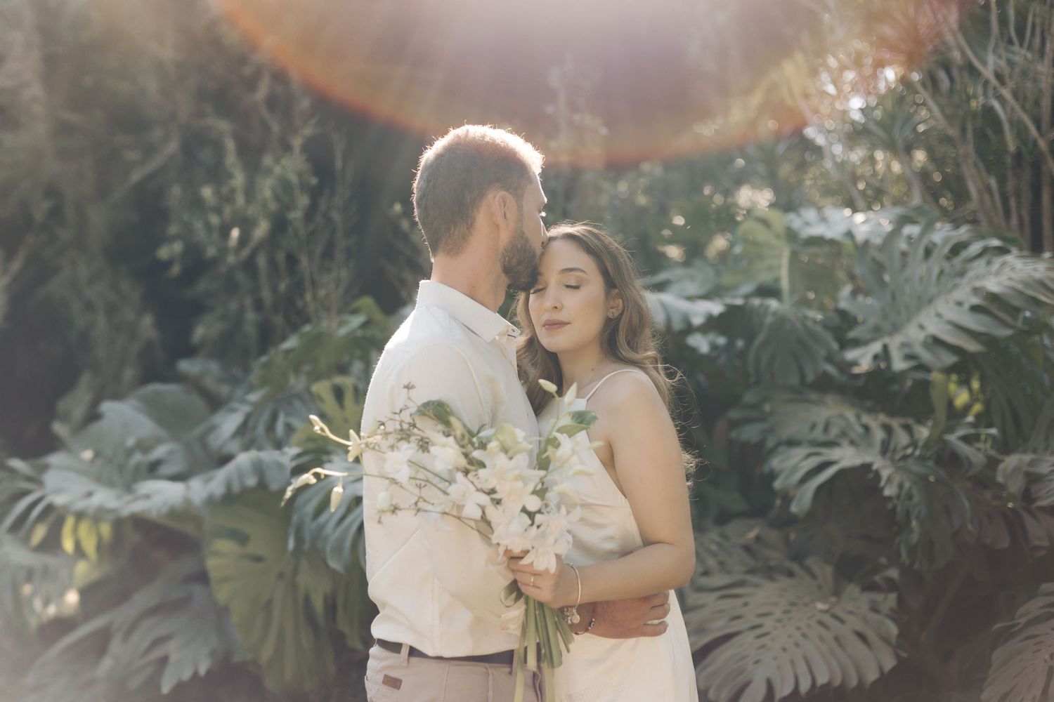 A couple shares an intimate moment in a tropical garden setting with light flares and white floral bouquet.