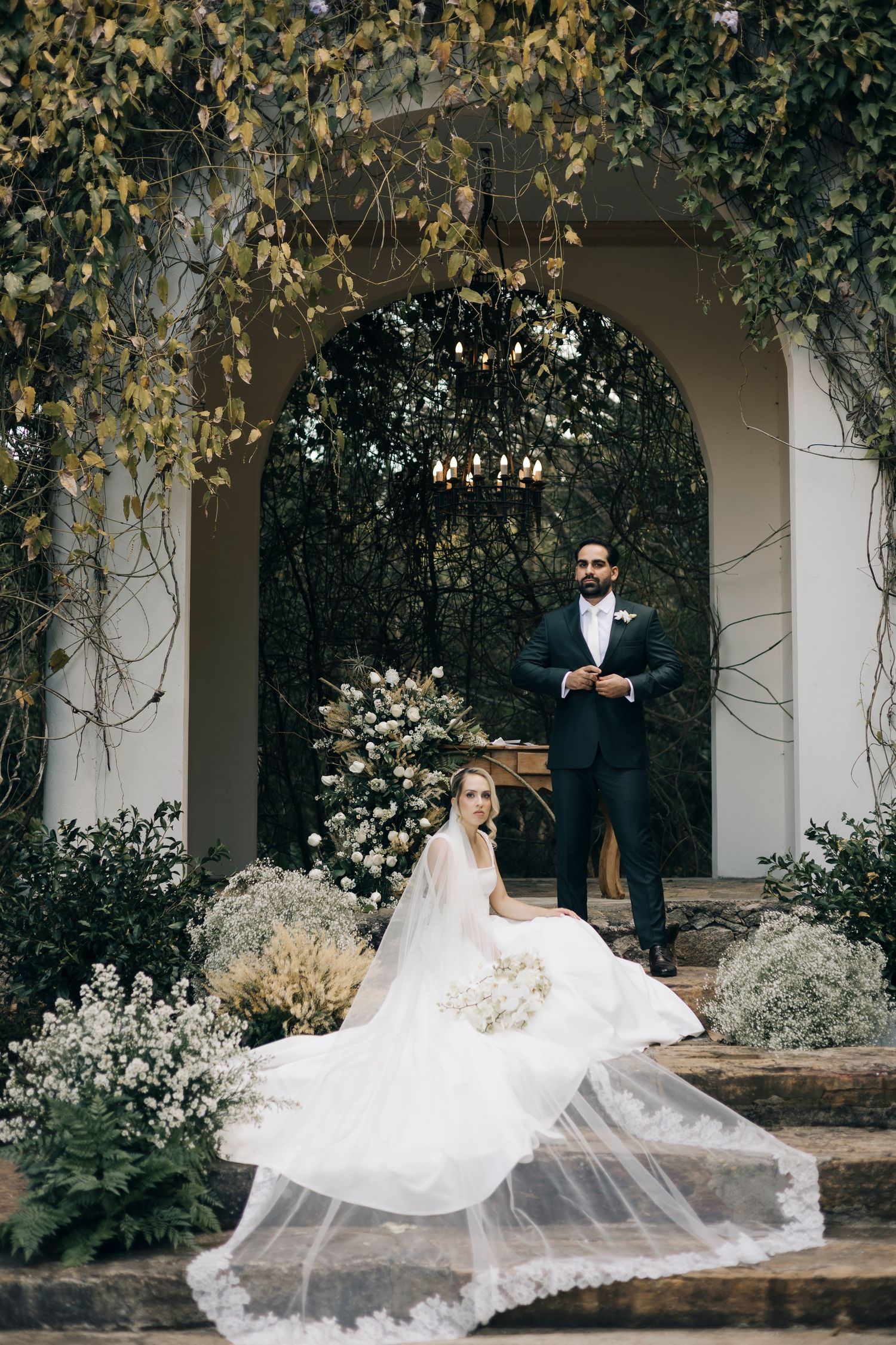 An elegant wedding couple poses on stone steps beneath an ivy-covered garden archway.