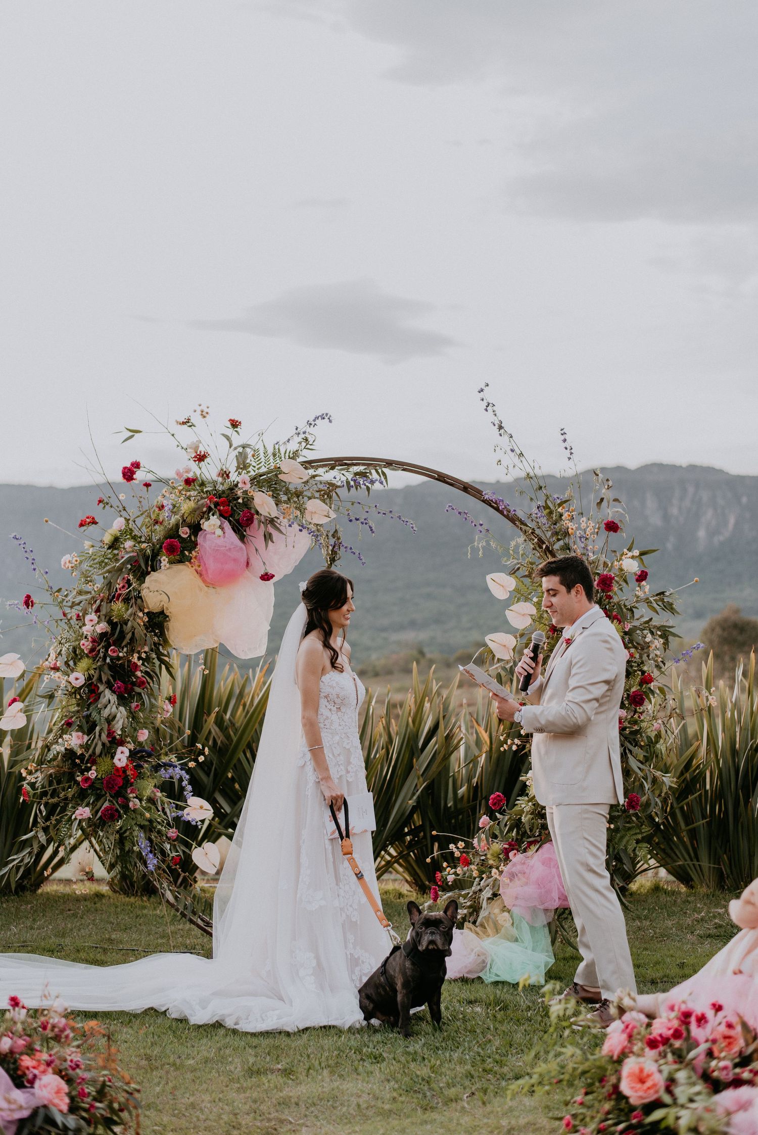 Intimate wedding moment under circular flower arch with pink balloons and mountain vista.