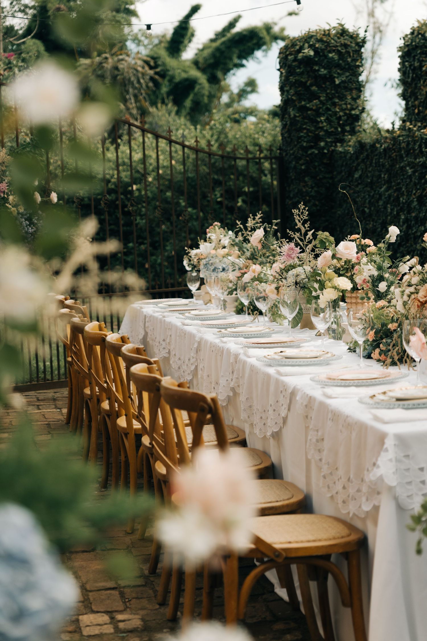 Elegant garden wedding table setting with white lace tablecloth and wooden chairs against lush greenery.